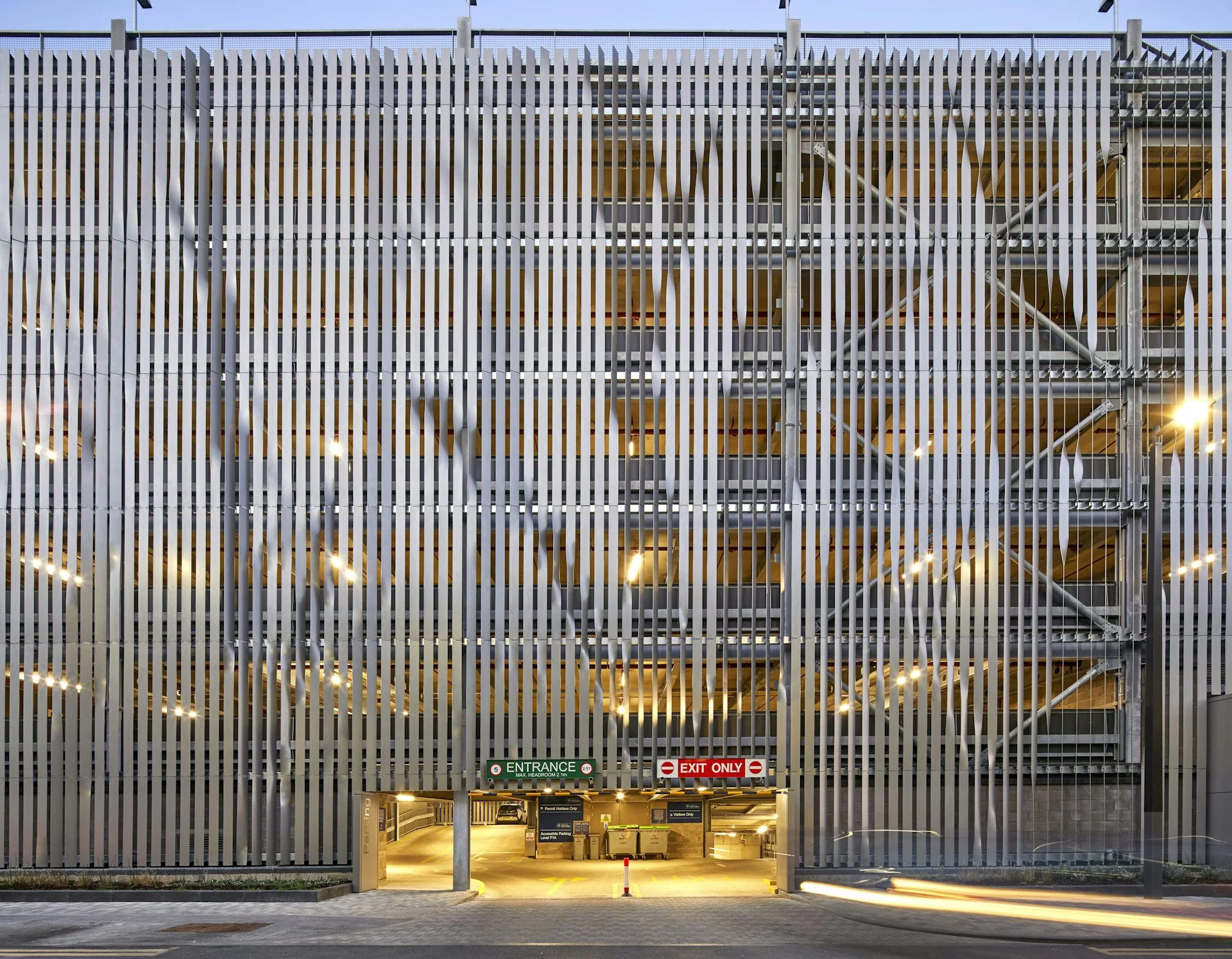 Four-story parking garage with a metal slat facade. Entrance and exit signs visible at ground level, with vehicles parked inside. Bright lights illuminate the interior.