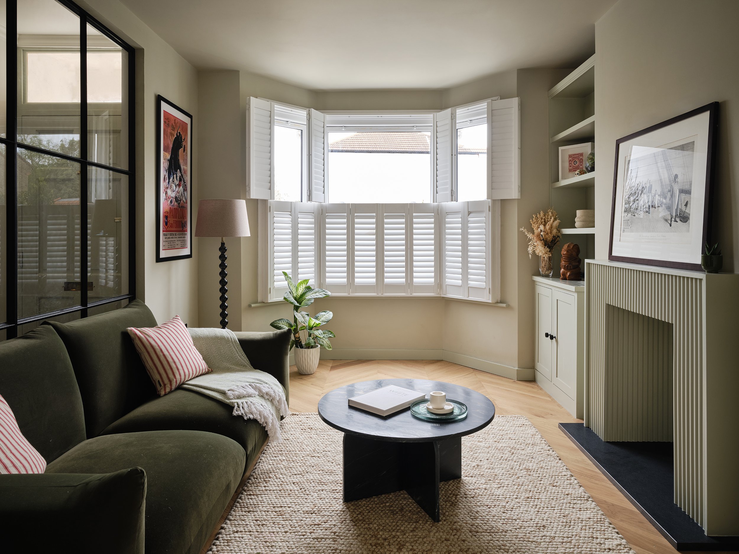 Living room with a large window with white shutters, a couch with striped pillows, a round coffee table, a floor lamp, framed artwork, and built-in shelves with decorative items.