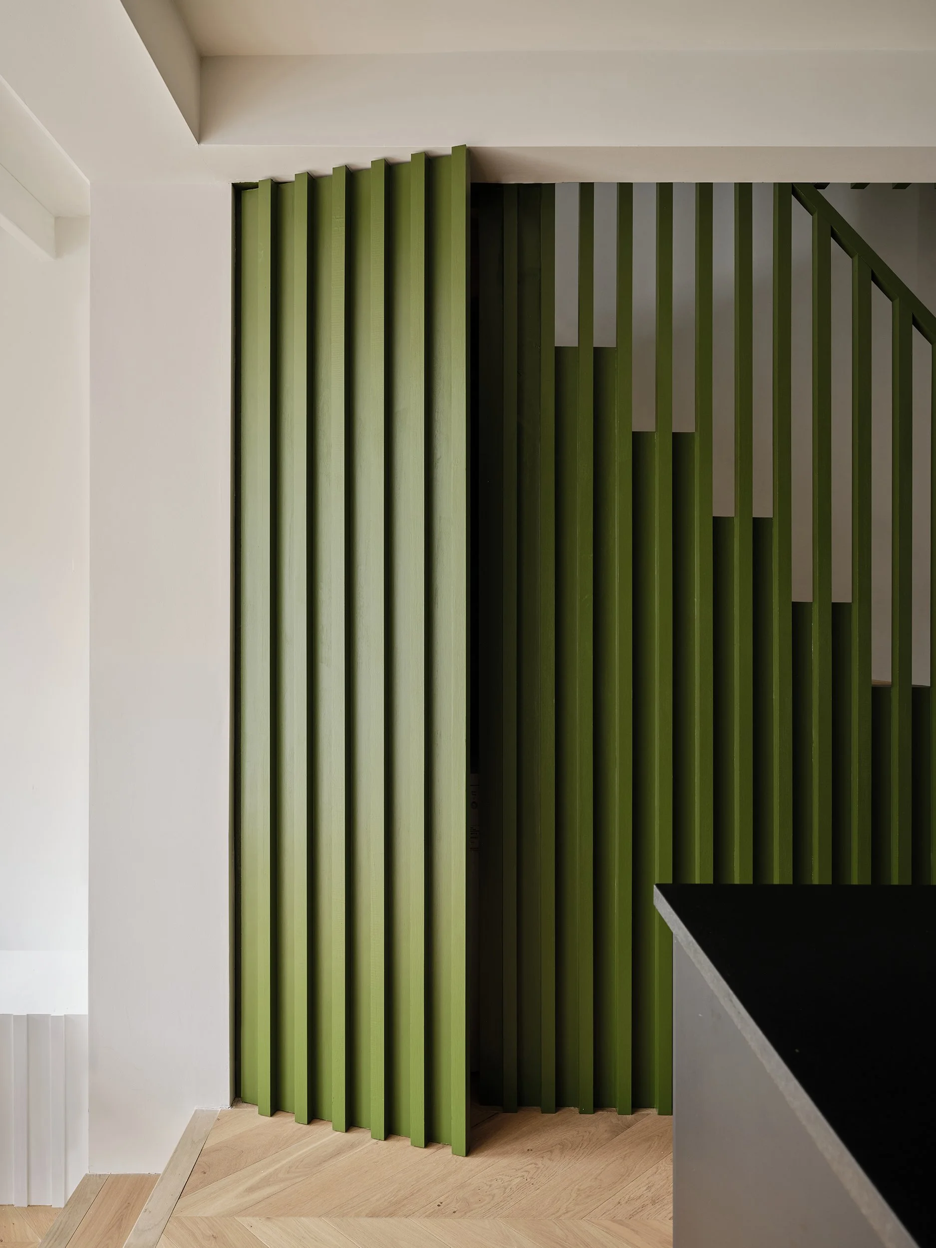 Interior view of a modern room with green vertical wooden divider panels, white walls, light wood flooring, and a black countertop on the right side.