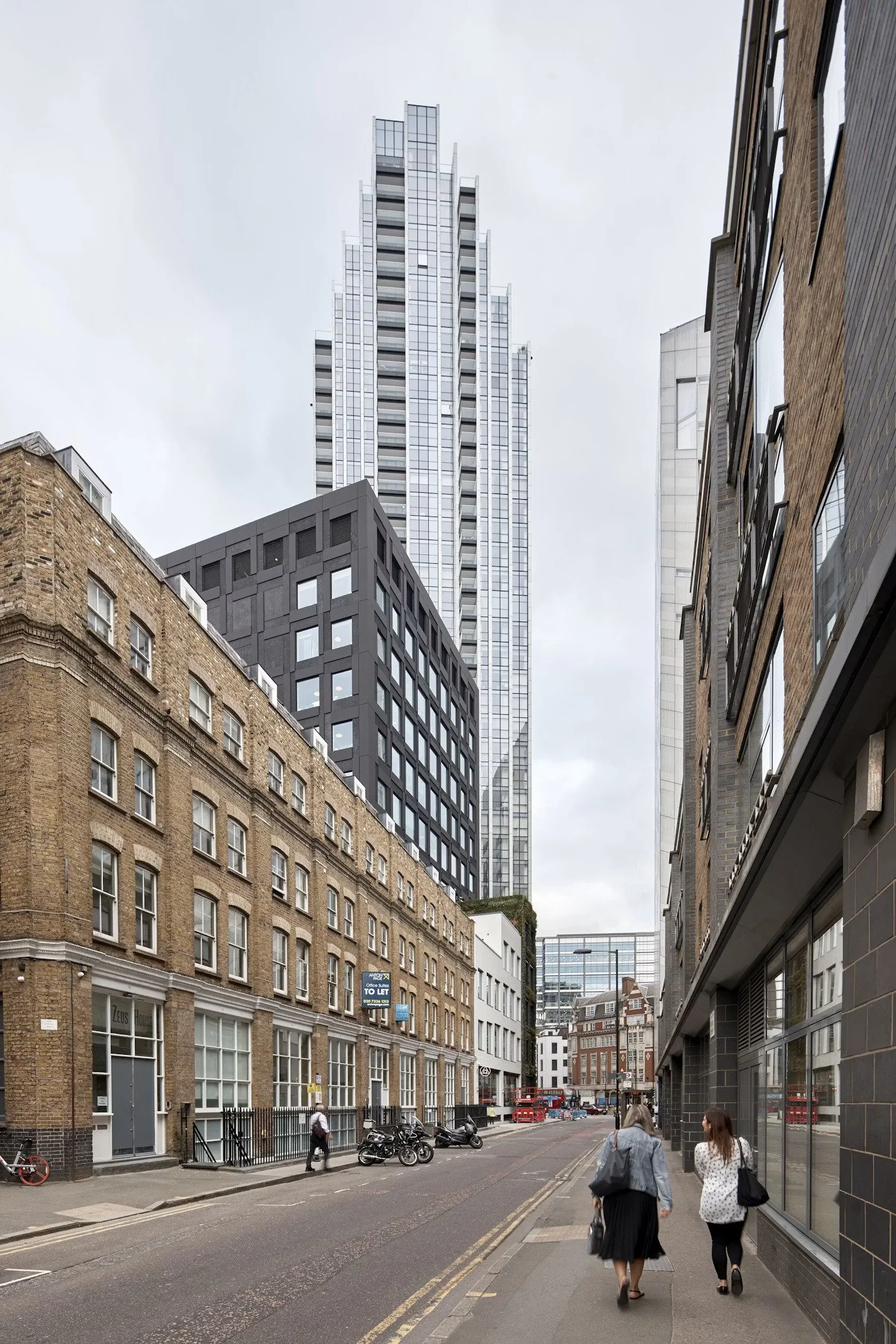 City street scene with multi-story buildings, pedestrians walking on sidewalk, bicycles and motorcycles parked, overcast sky.