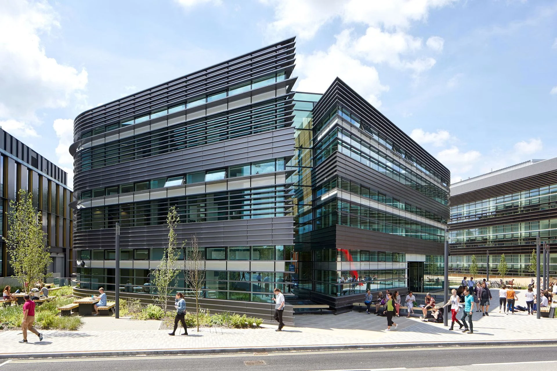 Modern multi-story office building with glass windows and sleek black horizontal paneling, situated in an urban area with pedestrians and outdoor seating in the foreground, under a partly cloudy sky.
