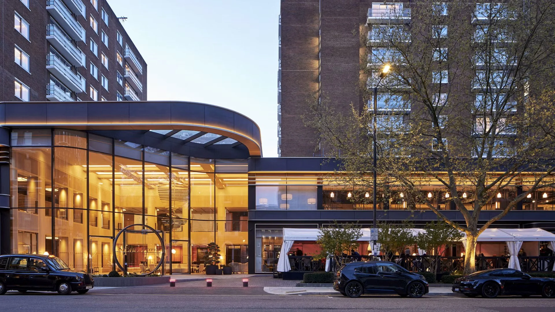 Modern building with large glass windows and warm interior lighting, surrounded by parked cars and trees in an urban setting at dusk.