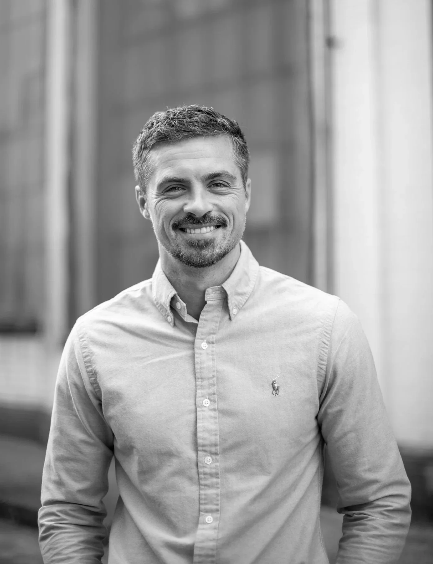 Black and white photo of a man with short hair, beard, and mustache, smiling, wearing a light-colored button-up shirt with a small logo on the left chest.