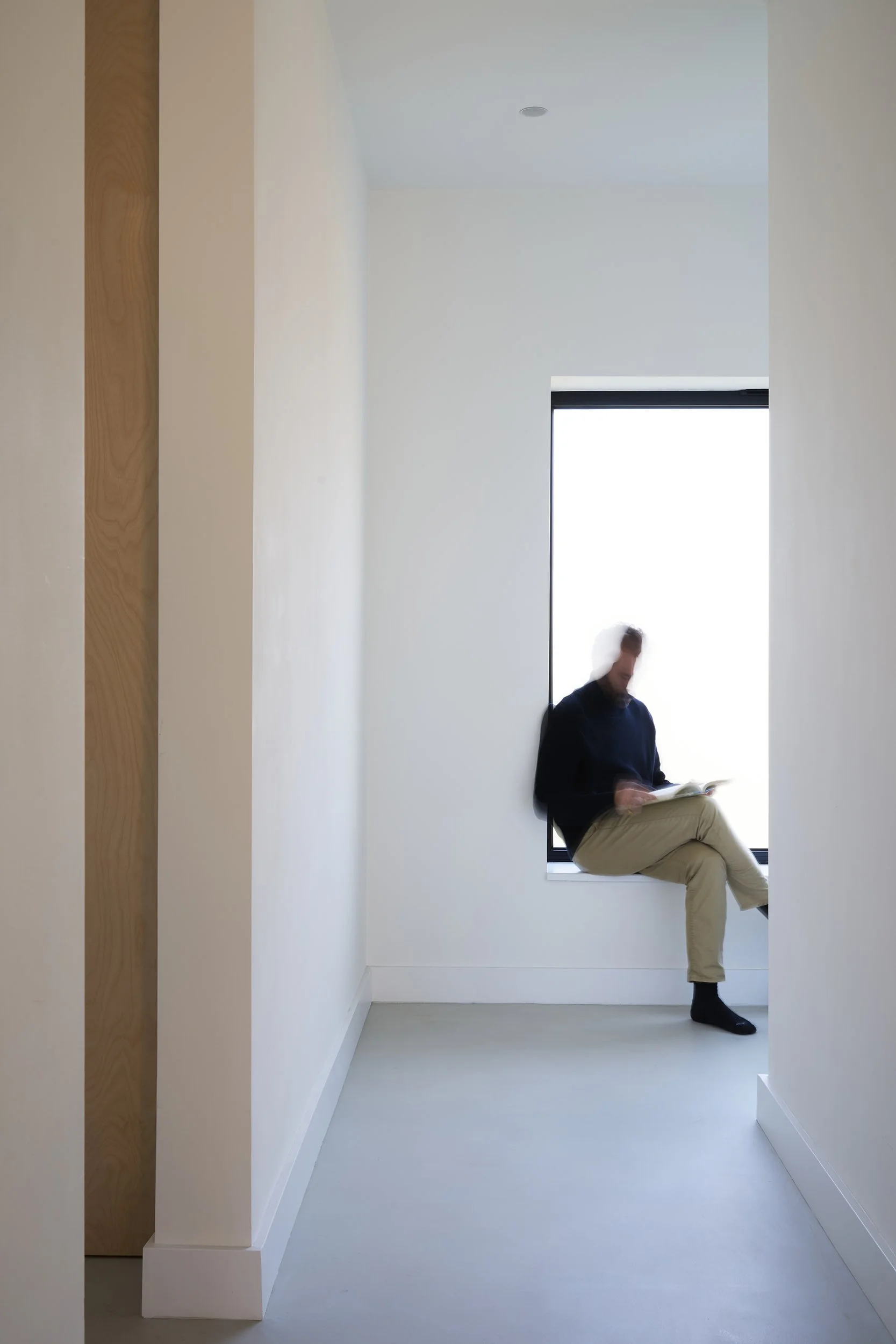 A man with dark hair wearing a dark sweater and khaki pants is sitting on a window ledge inside a minimalistic, white room, reading a book. The scene includes plain white walls, a large window, and light-colored flooring.
