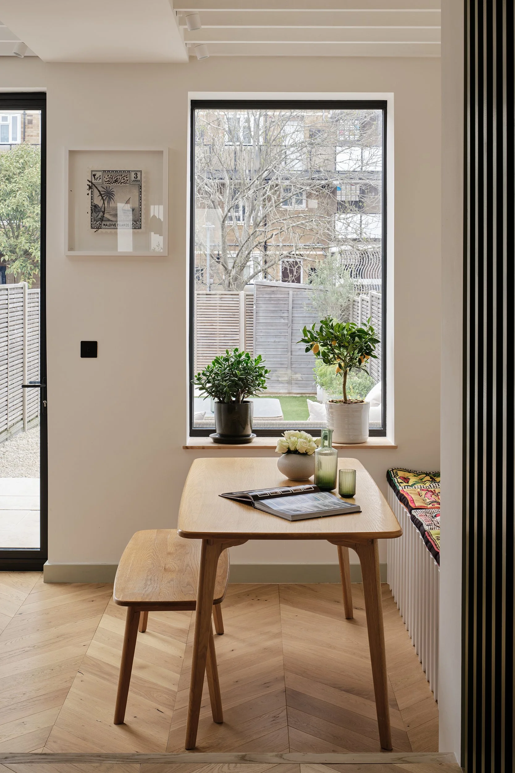 A dining area with a wooden table and matching bench, a window with plants on the sill, and outside view of trees and neighboring buildings.