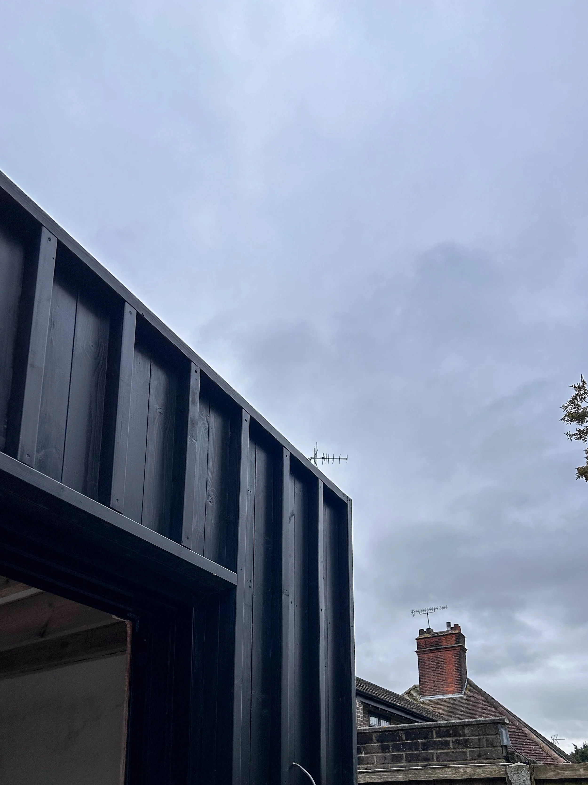 Part of a black house with siding, an open garage door, a chimney, and rooftops against a cloudy sky.