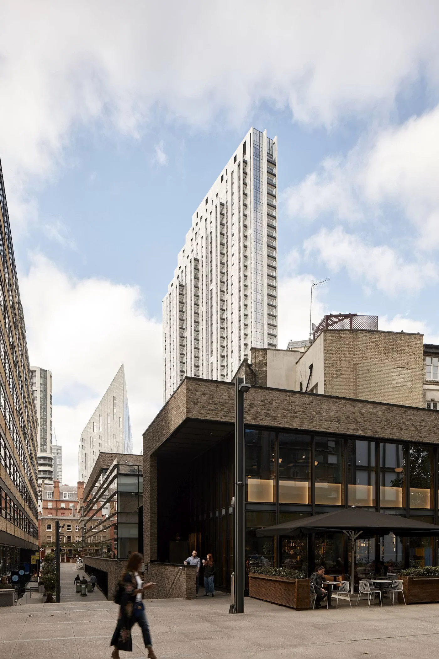 Urban scene with modern skyscrapers, an outdoor dining area with tables and chairs, and people walking on a sidewalk.