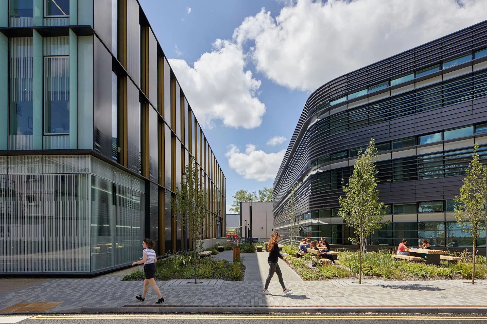 Modern office buildings with glass and metal facades, a landscaped courtyard with seating and trees, and people walking and sitting outdoors on a sunny day.