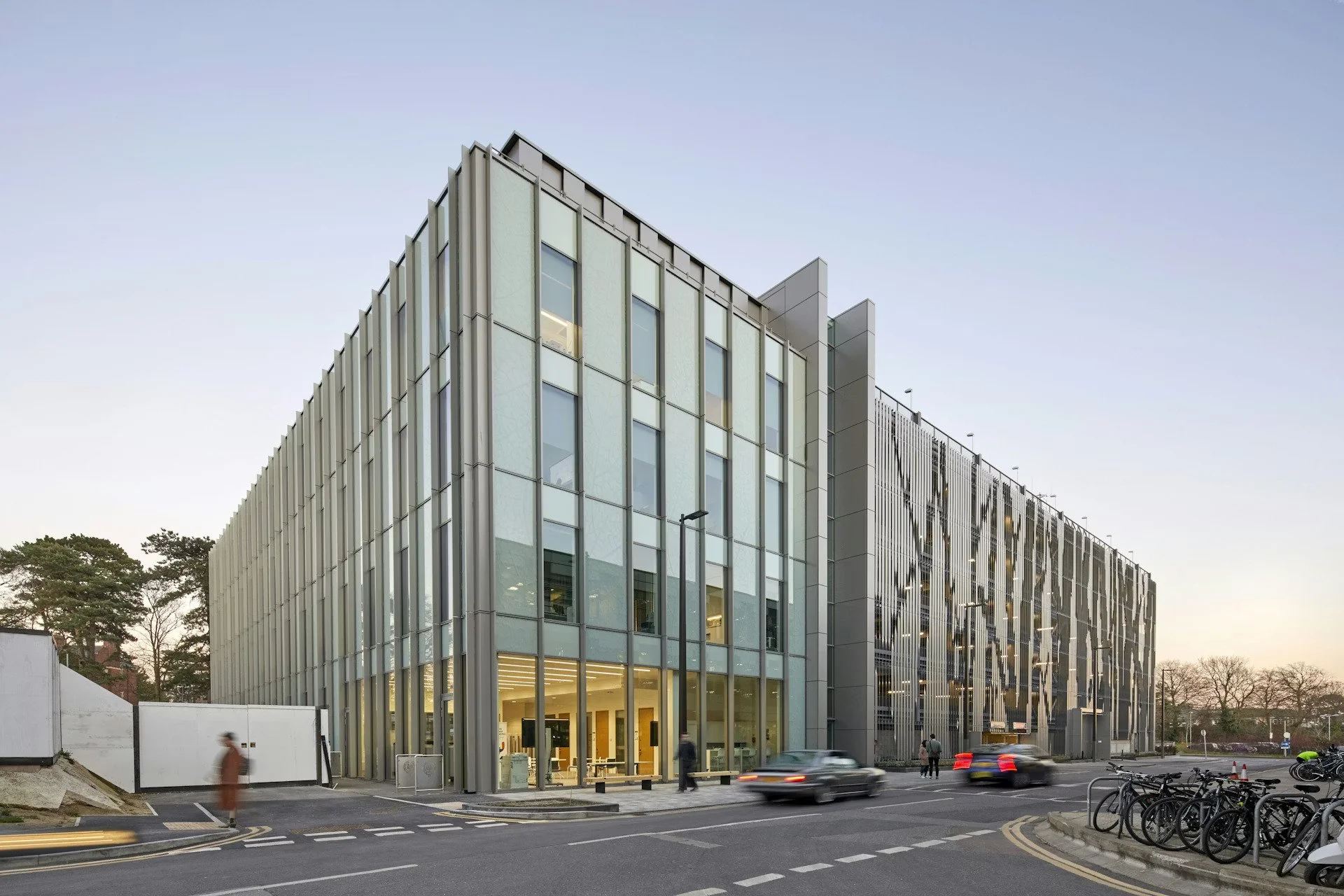 Modern glass building with vertical metal panels, situated along a city street with cars, bicycles, and pedestrians, during sunset.