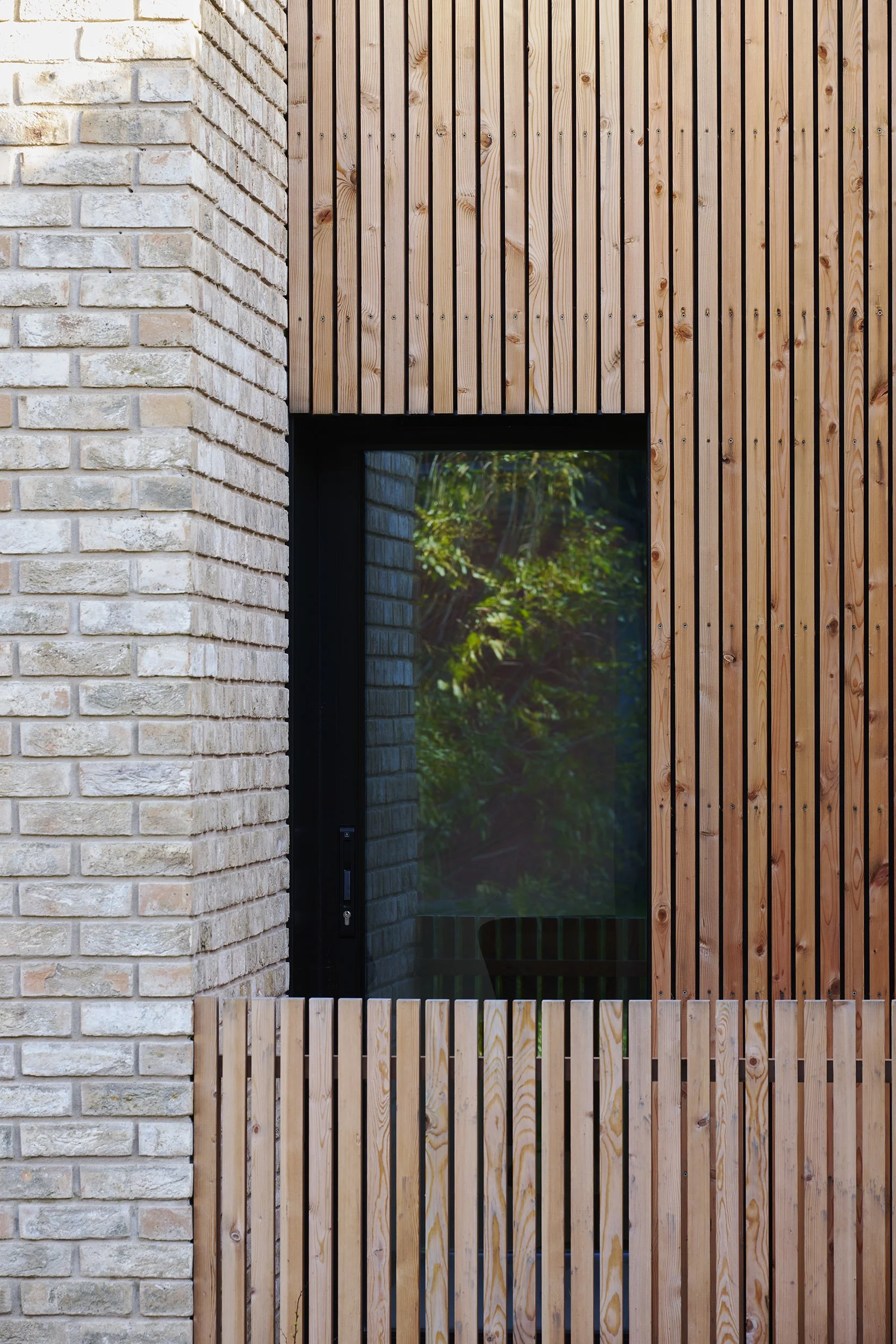 Exterior view of a modern building with brick wall on the left and wooden paneling on the right, featuring a window with greenery reflected in it.