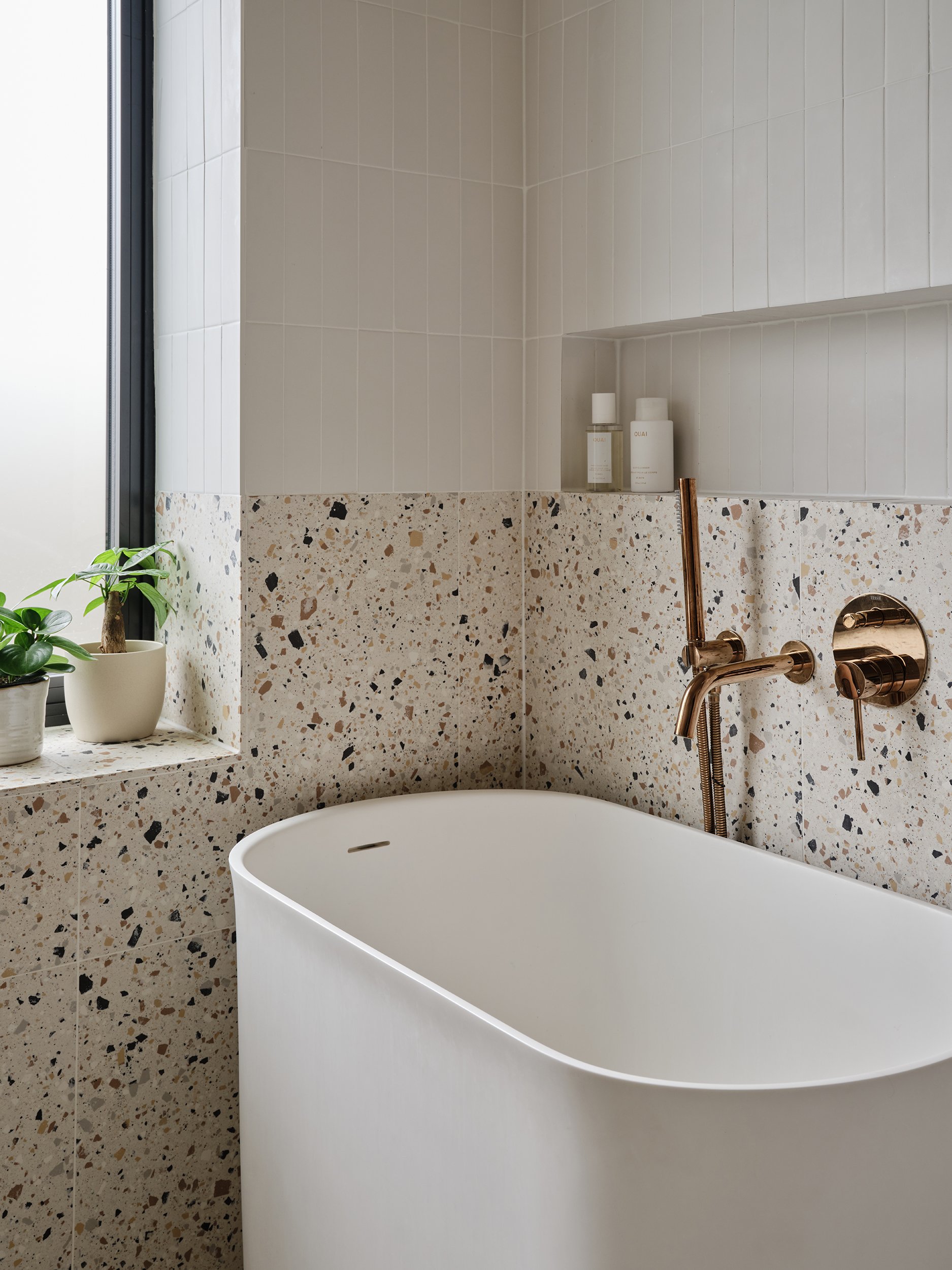 A modern bathroom features a white freestanding bathtub with a rounded design, copper fixtures, and a terrazzo countertop with potted plants on a ledge. The wall above has white subway tiles, and bottles of bathroom products sit in a niche. A window 