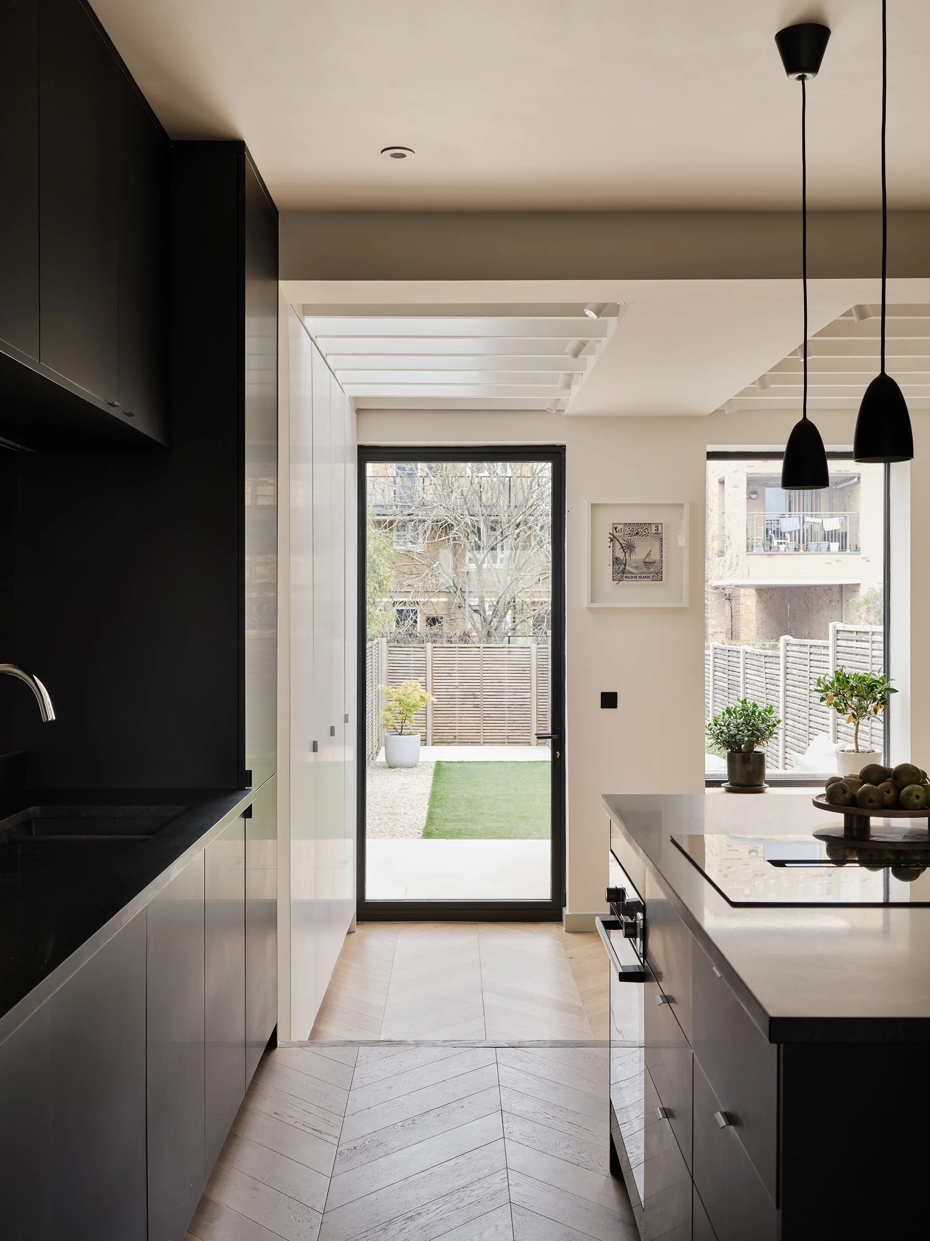 Modern kitchen with black and white cabinetry, a countertop with a bowl of apples, two black pendant lights, large windows showing an outdoor patio, small potted plants, and a wooden fence.