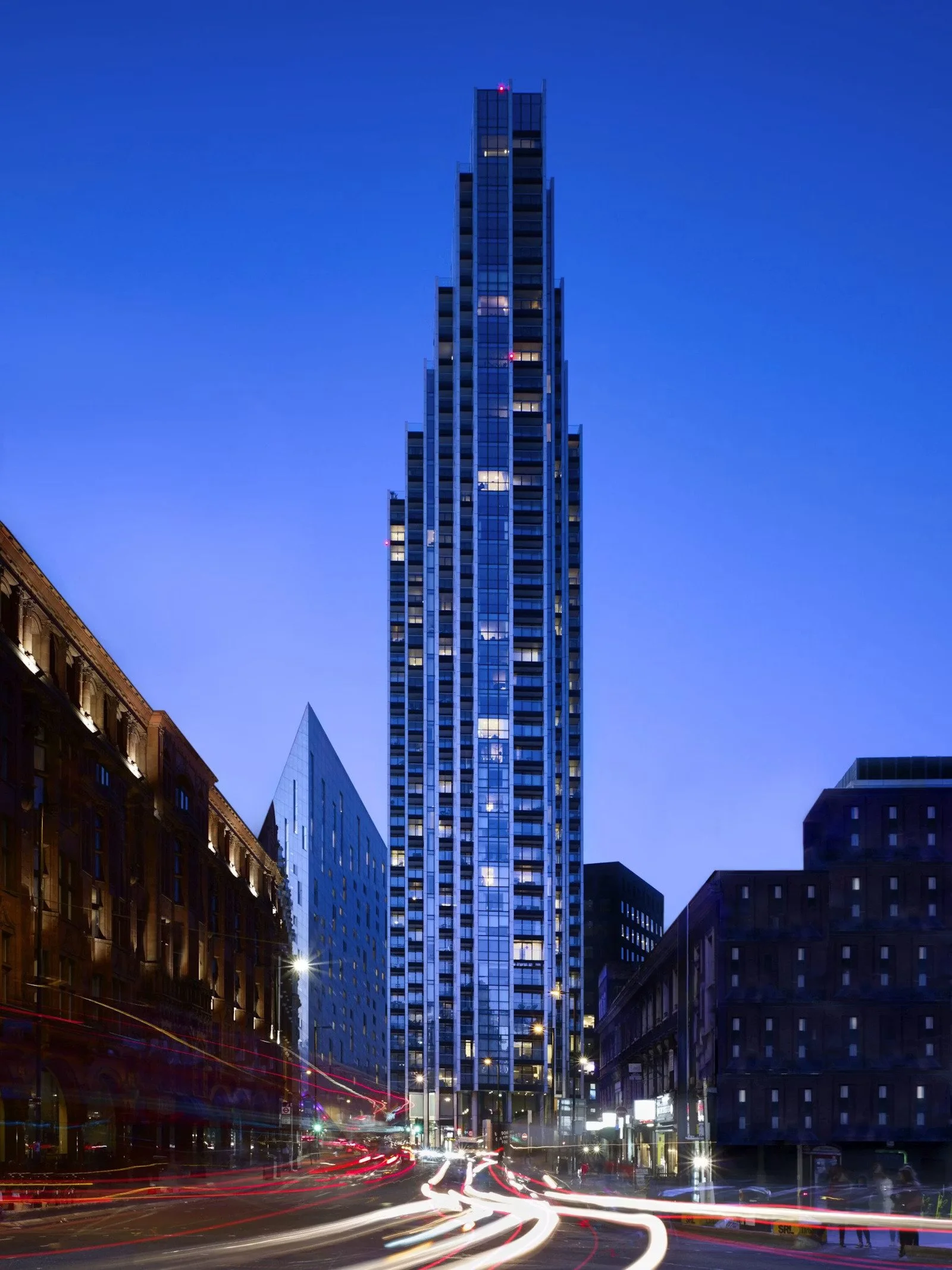 Tall modern skyscraper illuminated against a deep blue evening sky, surrounded by shorter buildings and bustling city streets with moving car light trails.