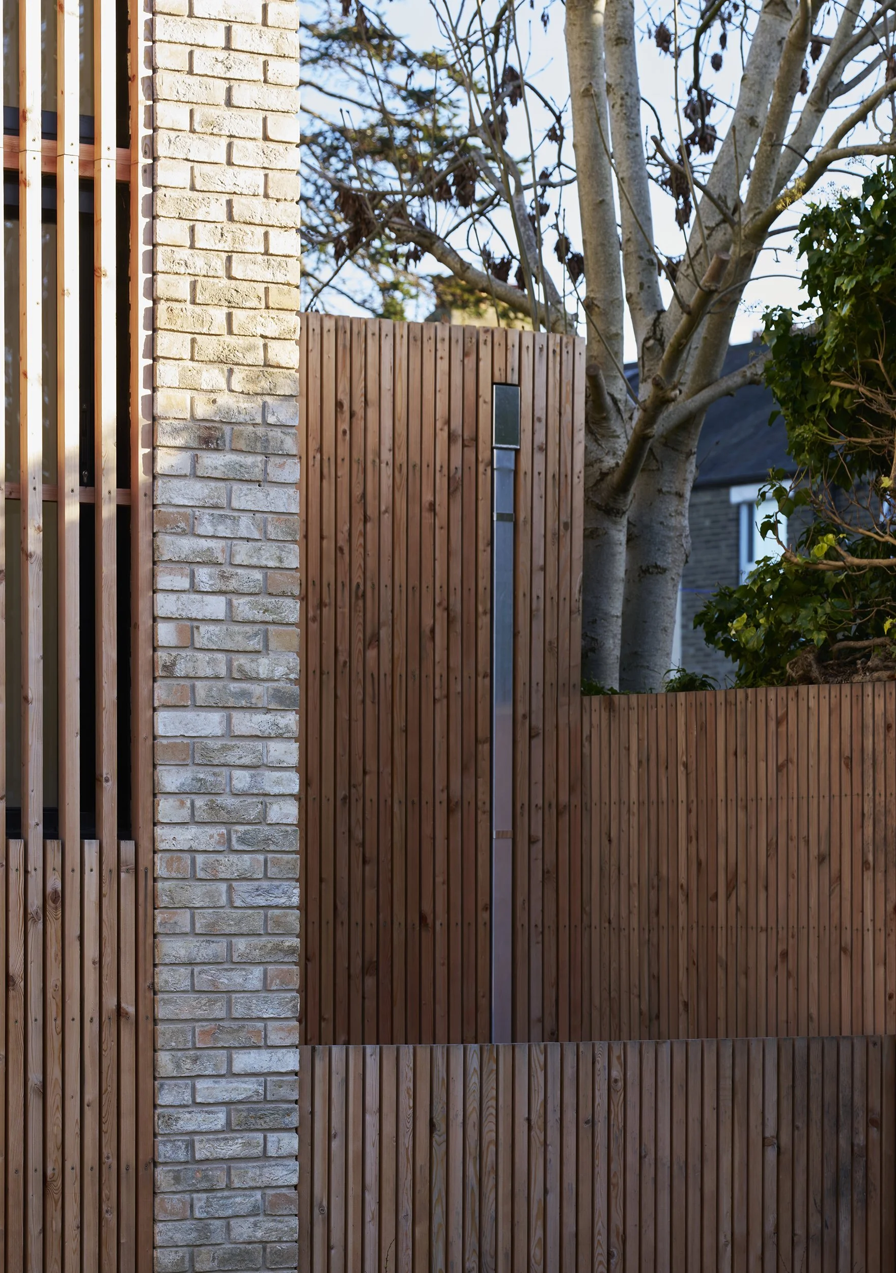 A modern outdoor wooden fence with vertical slats, a brick pillar on the left, and a tree with bare branches and some green foliage behind the fence. A small blackbox is mounted on the fence.