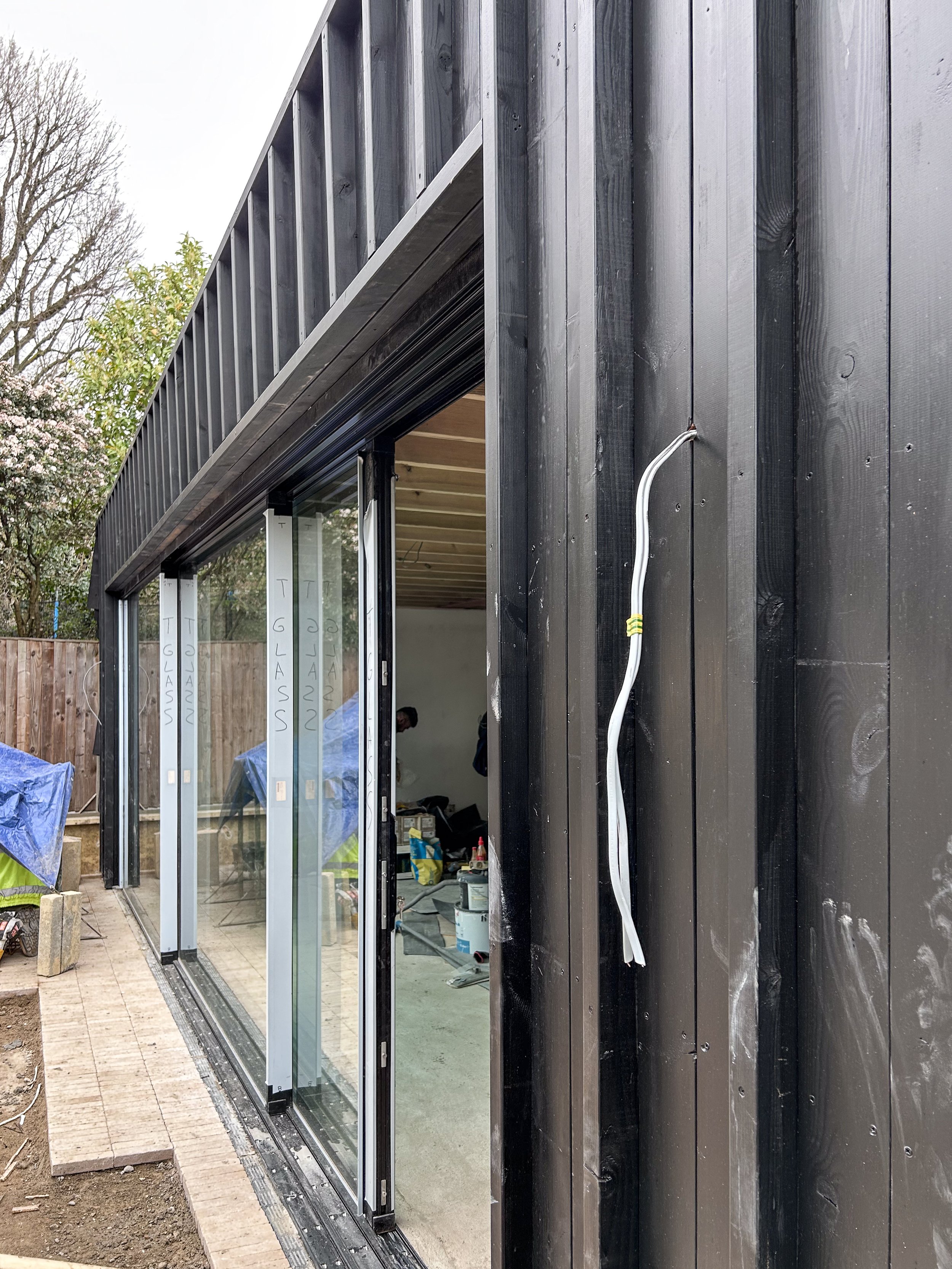 Exterior view of a building under construction with large glass sliding door, black wooden siding, and construction tools and materials inside.