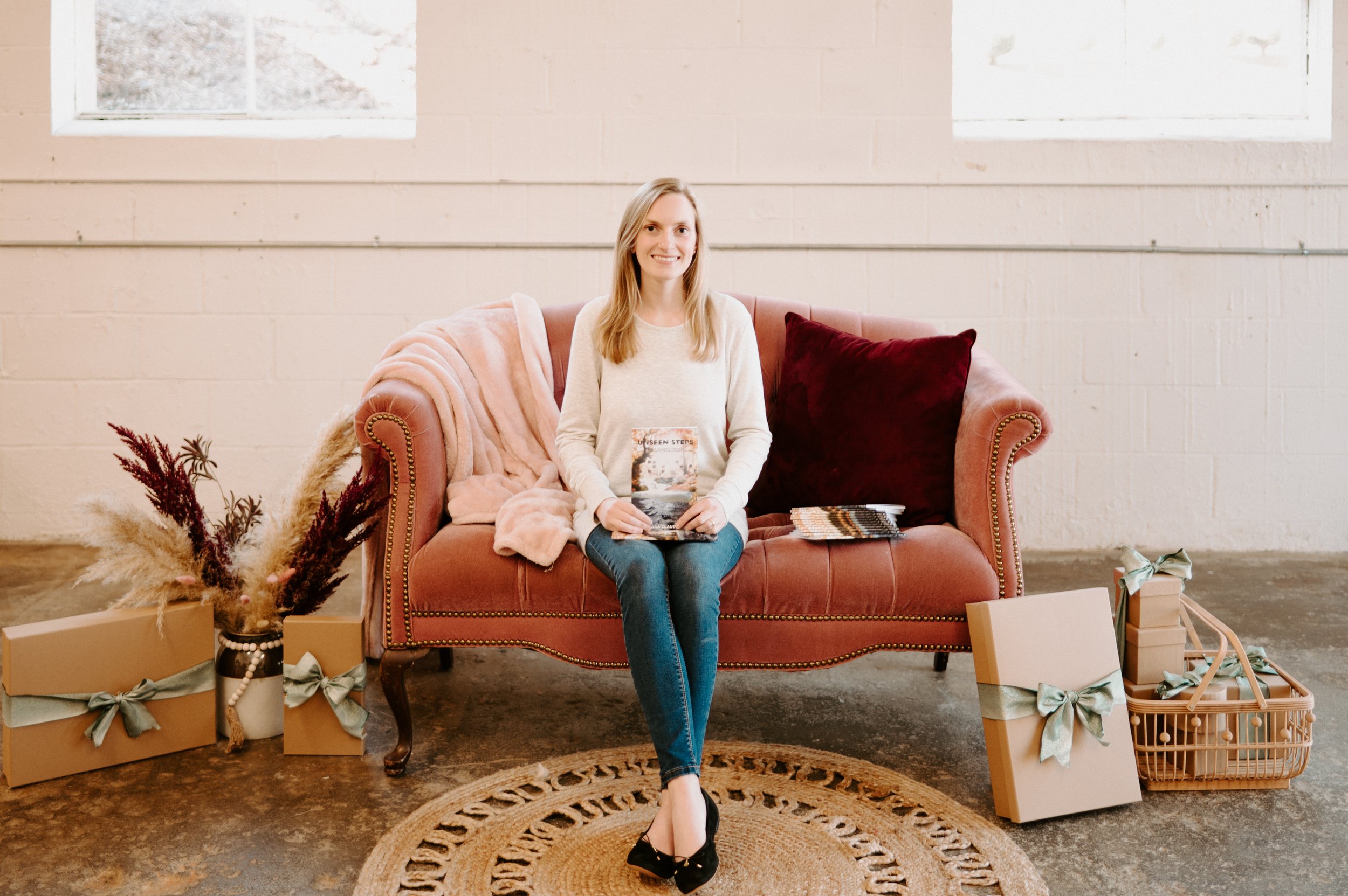 Full body shot of SC in cream top and jeans, sitting on couch with ankles crossed, holding Unseen Steps in lap, smiling at the camera.jpg
