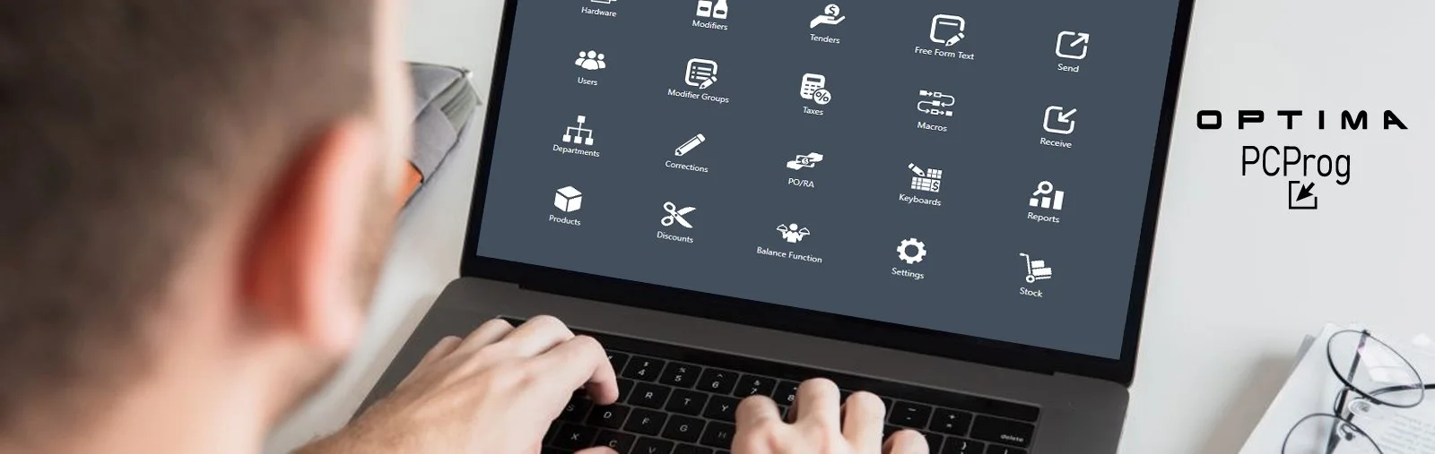 A person using a laptop with a screen displaying various software icons and labels, including 'Hardware,' 'Modifiers,' 'Tenders,' and others. The person's head and hands are partially visible, and there is a white surface with glasses and a coil-bound notebook nearby.