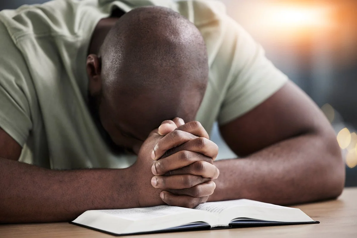 Close up image of a black man sitting at a desk with his scriptures opened. His hands are clentched tightly in prayer on top of the book and his head is bowed in prayer.