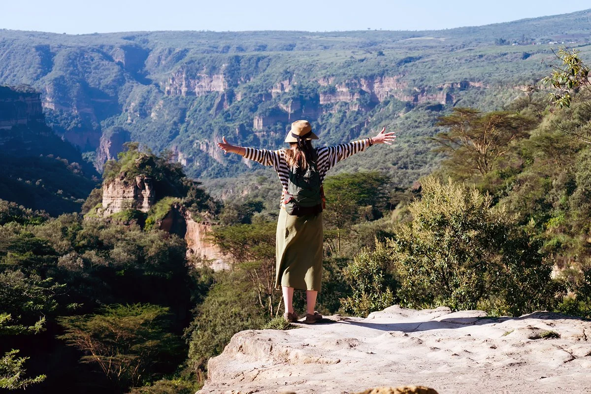 Woman travler standing on the edge of a rock that opens up to a vast view of mountains and greenery. Her arms are stretched out wide as if welcoming the expanse.