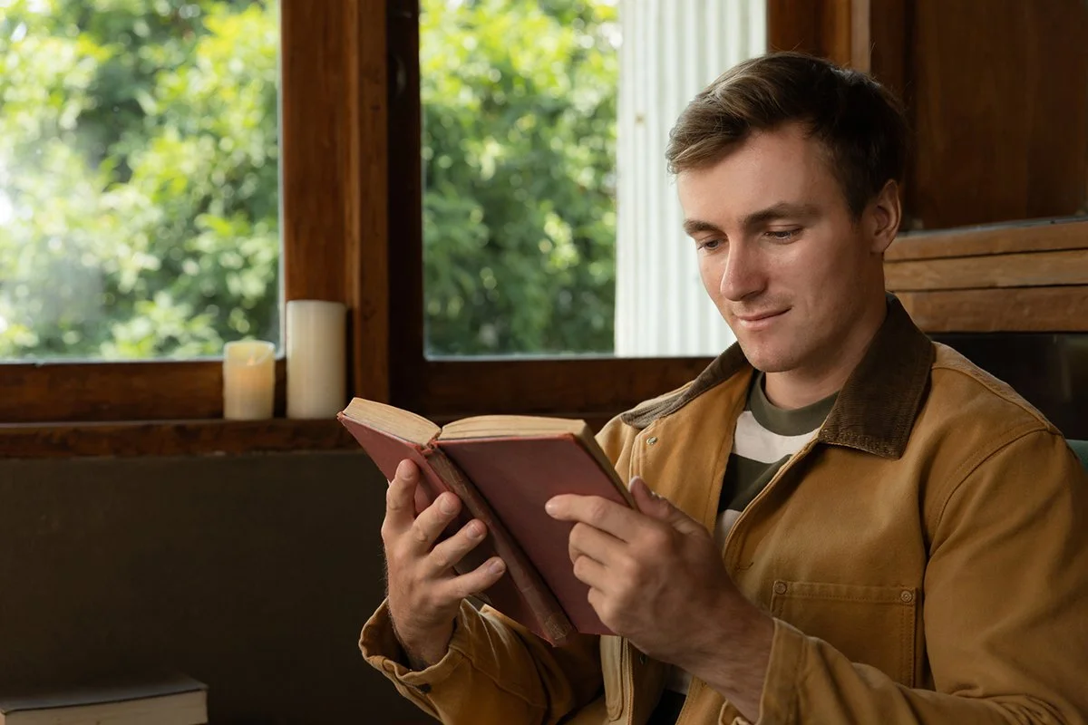 Young man sitting by a window reading an open book of scriptures.