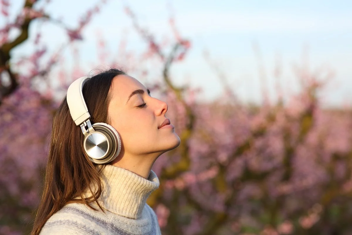Woman listening to headphones with eyes closed outside with spring trees in bloom in the background