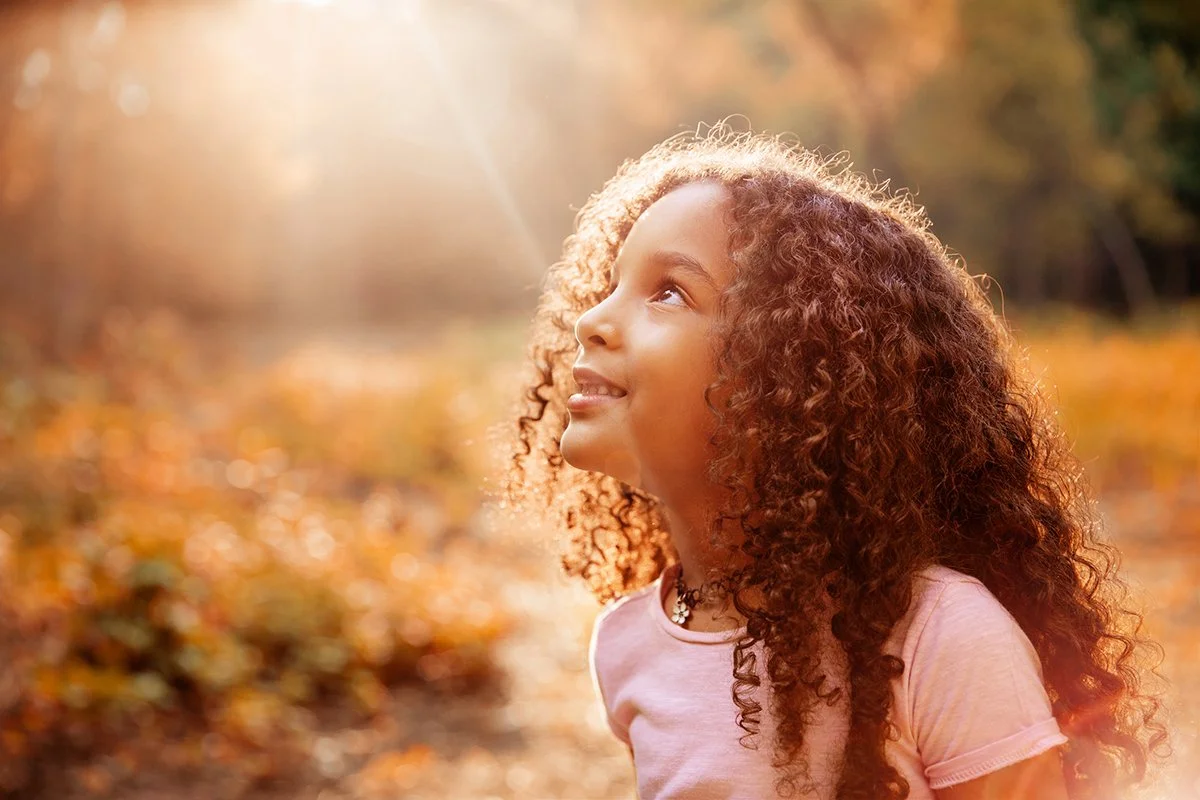 cute little girl with long curly hair, in nature, looking up, while receiving sun rays on her face