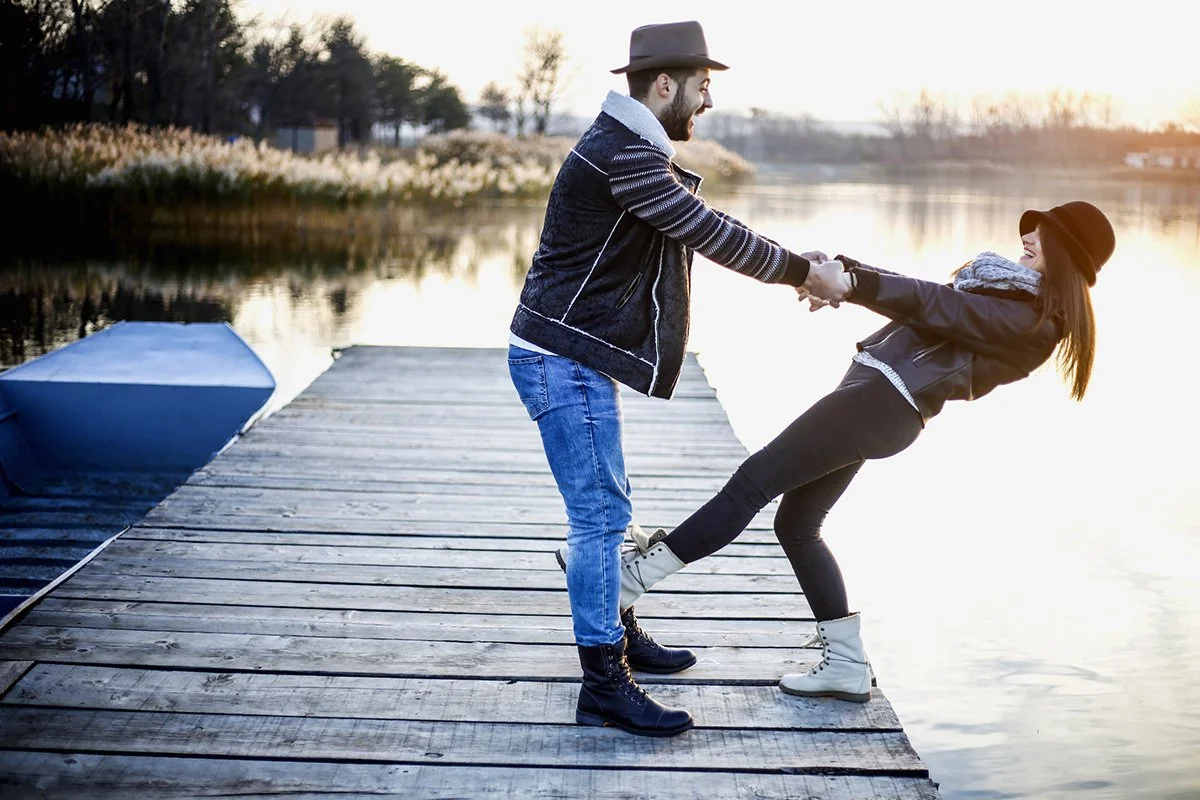 Couple on a pier, they are holding hands and the girl is leaning way back as if to fall in the water but the guys grip is keeping her from falling.