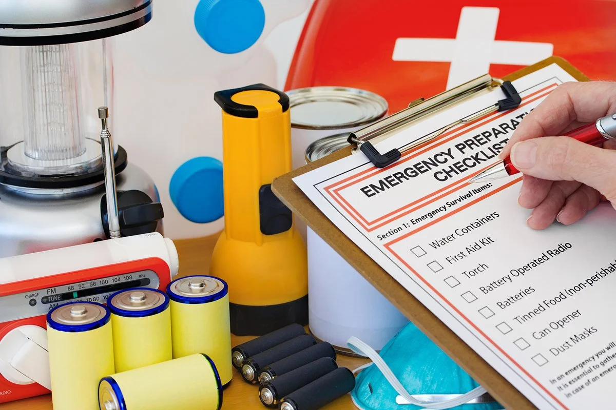 Photo of hand filling out an Emergency Preparation List, surounded by emergency items.