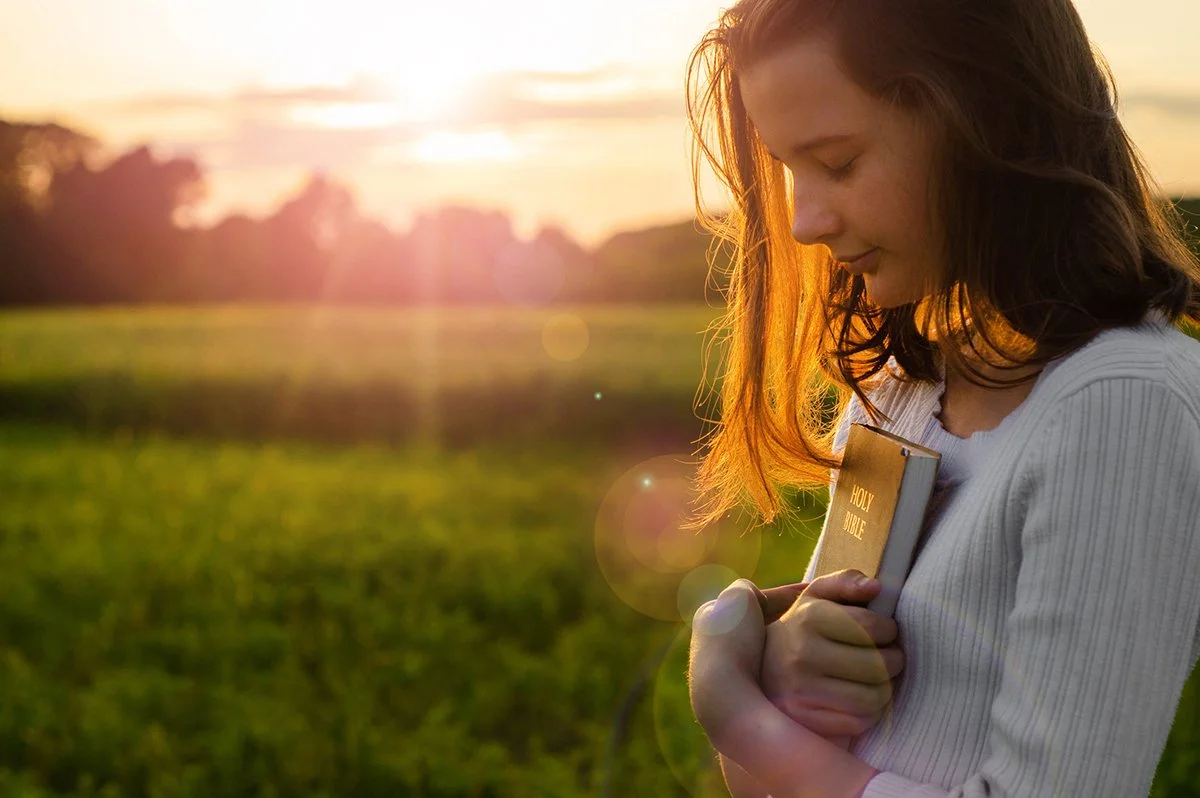 Young woman standing in a green field at sunset, clenching a bible to her chest with her head bowed and a peaceful smile.