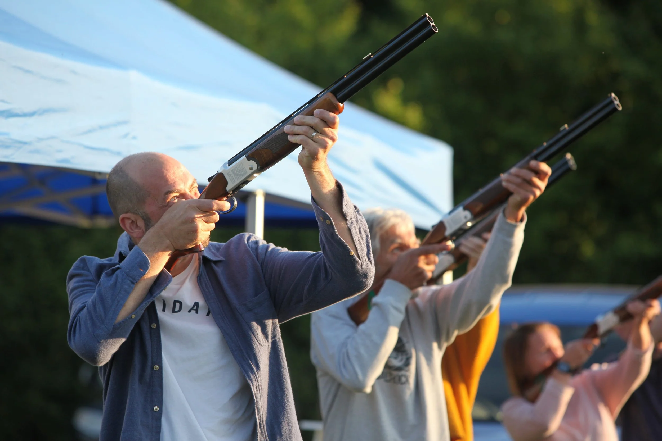 Group of people firing clay shotguns outdoors, standing side by side in a row with tents and trees in the background.