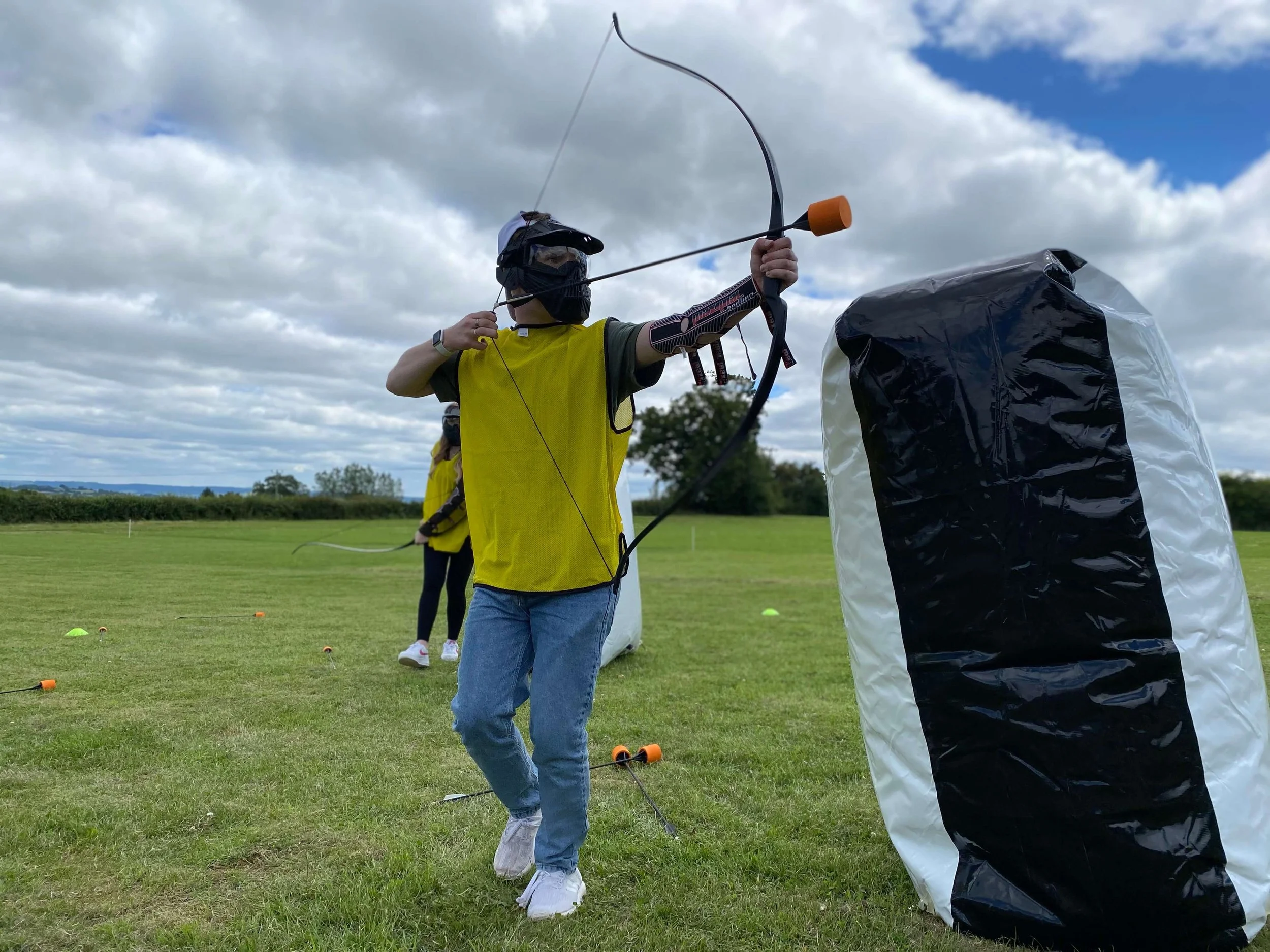 A person aiming a bow and arrow during archery tag outdoors on a cloudy day, with other participants in the background.