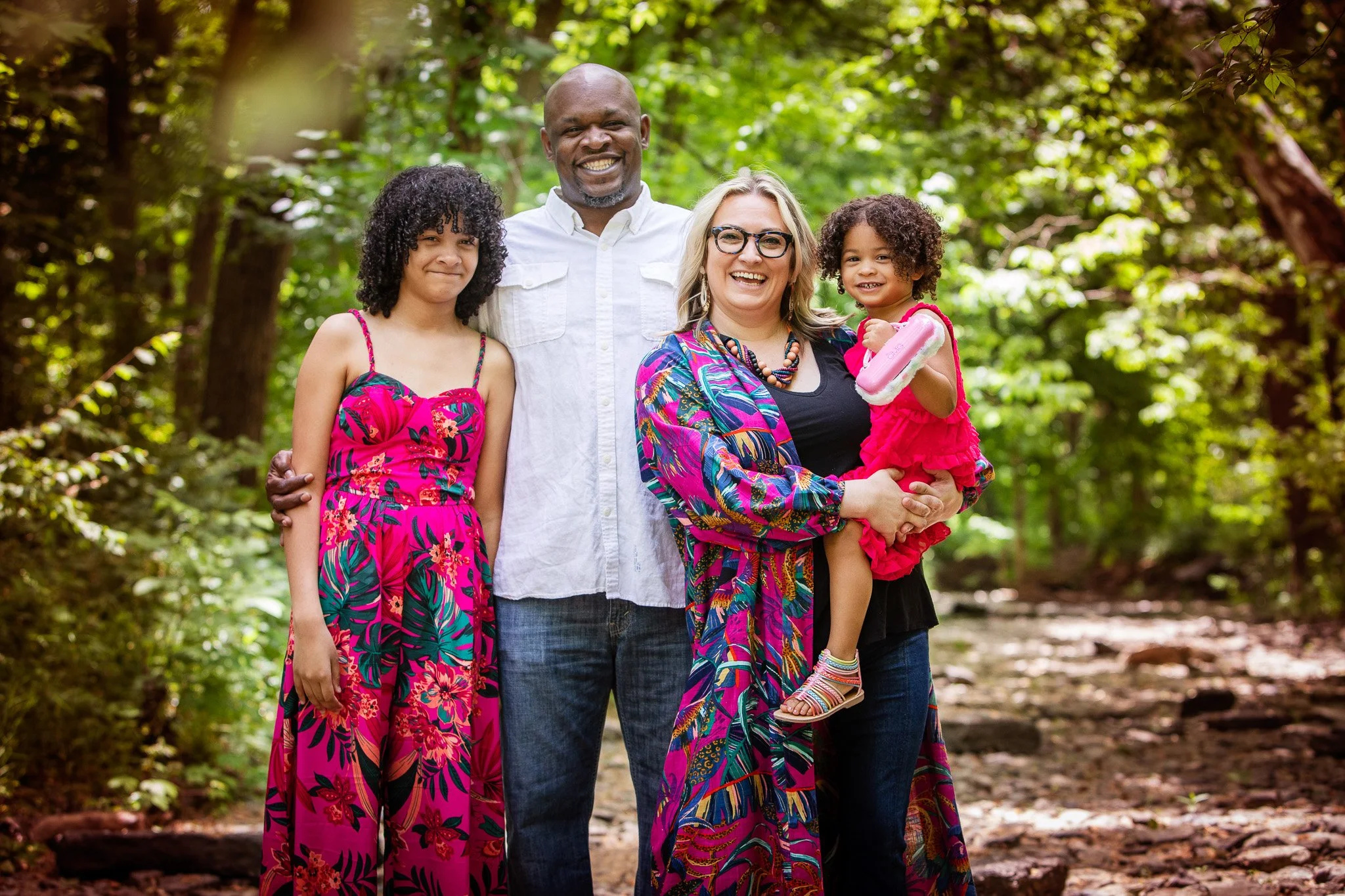 A multiracial family of four standing together outdoors in a green, wooded area, all smiling and looking at the camera. The woman on the right is holding a young girl dressed in pink, and the man behind her has his arm around the girl on the left. Th