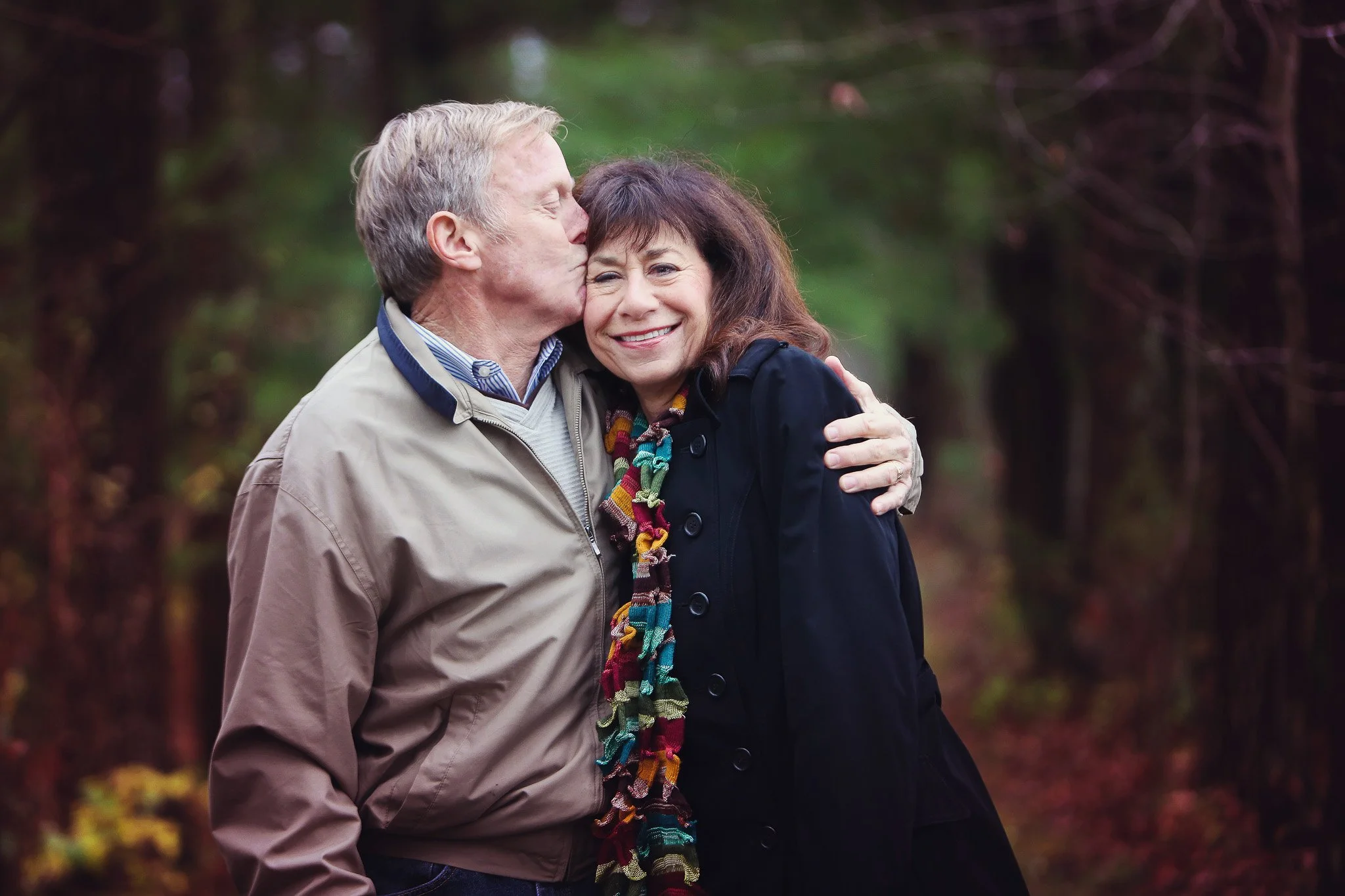 An elderly man kissing an elderly woman on the cheek while hugging her in a forested area.
