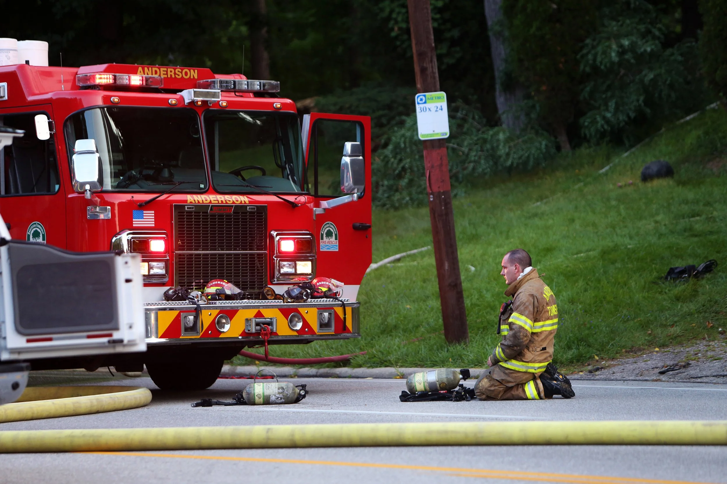 A firefighter kneeling on the road near a fire truck with emergency lights, surrounded by hoses and oxygen tanks.
