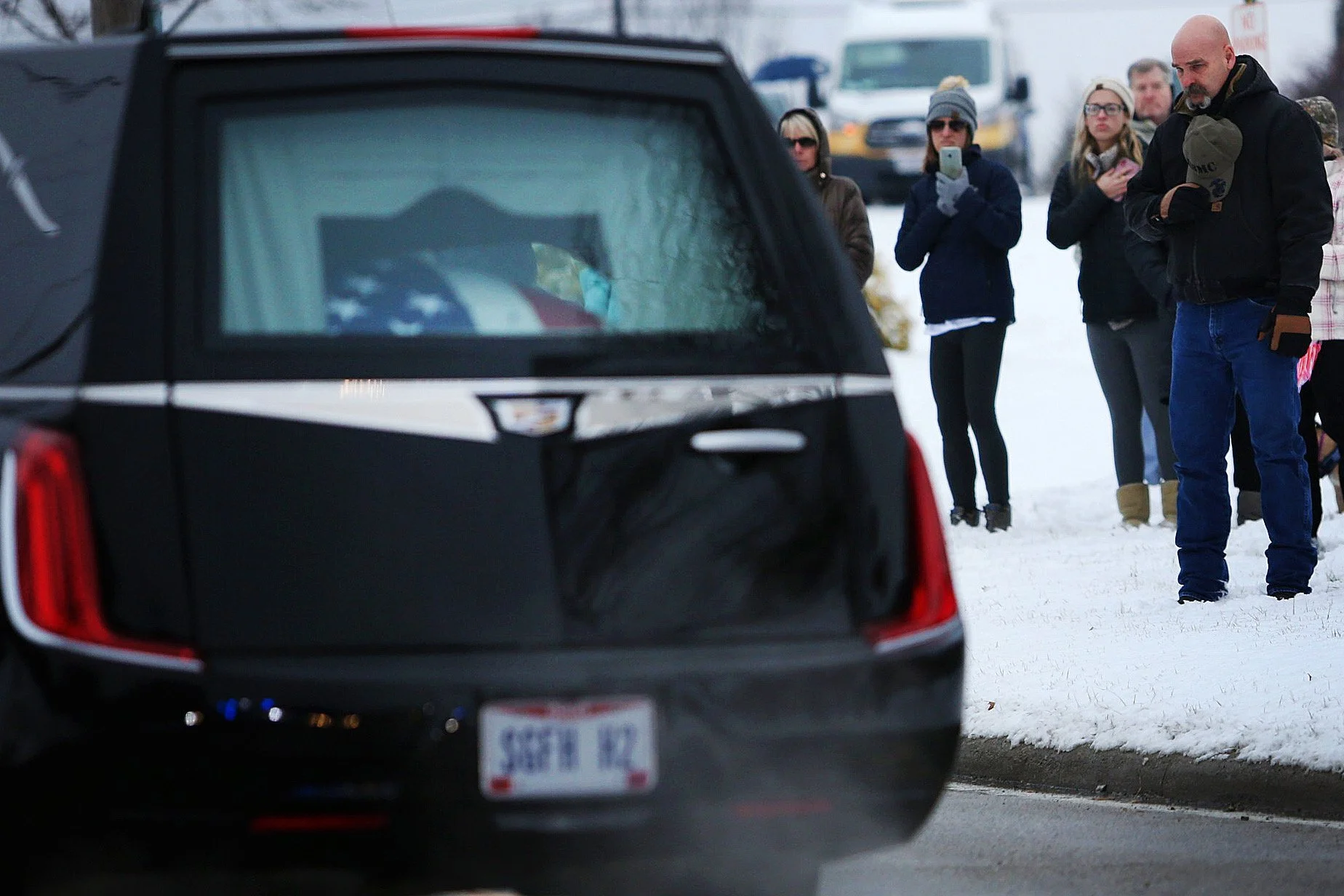 People standing on snowy ground, taking photos or observing a scene, with a black emergency vehicle in the foreground.