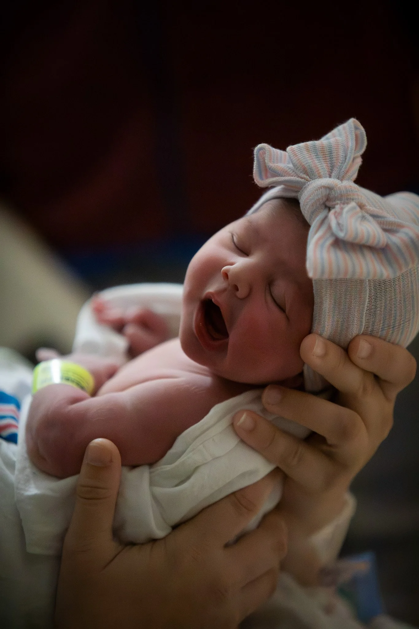 A newborn baby with a pink and blue striped hat is being held and gently supported by an adult's hands, yawning with eyes closed, and wrapped in a white blanket.
