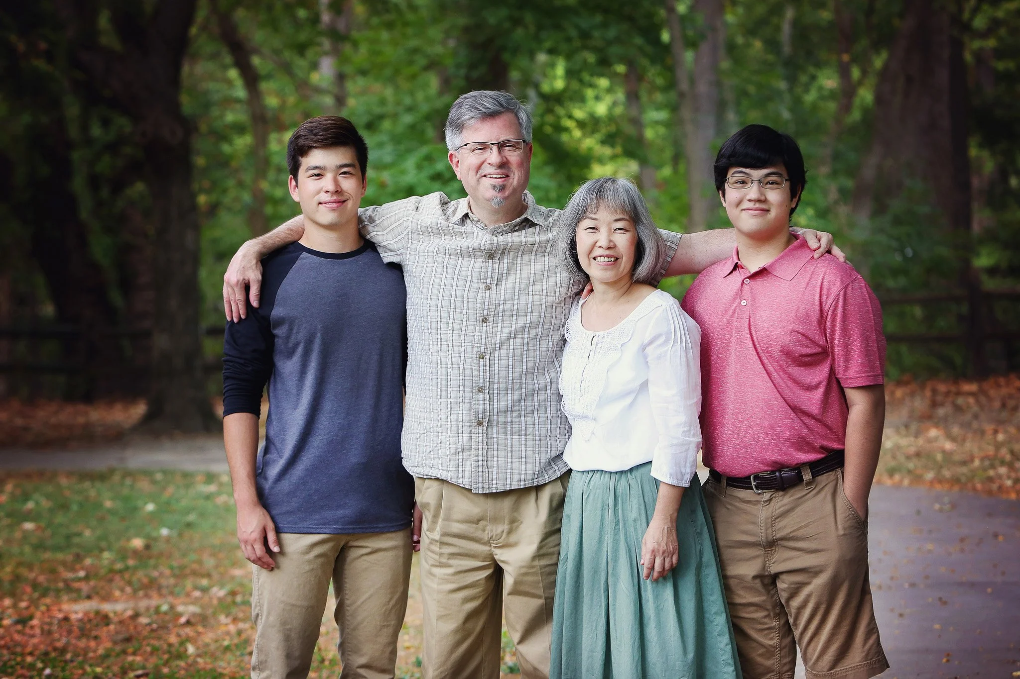 A multigenerational family of five standing outdoors in a wooded park, smiling with arms around each other.