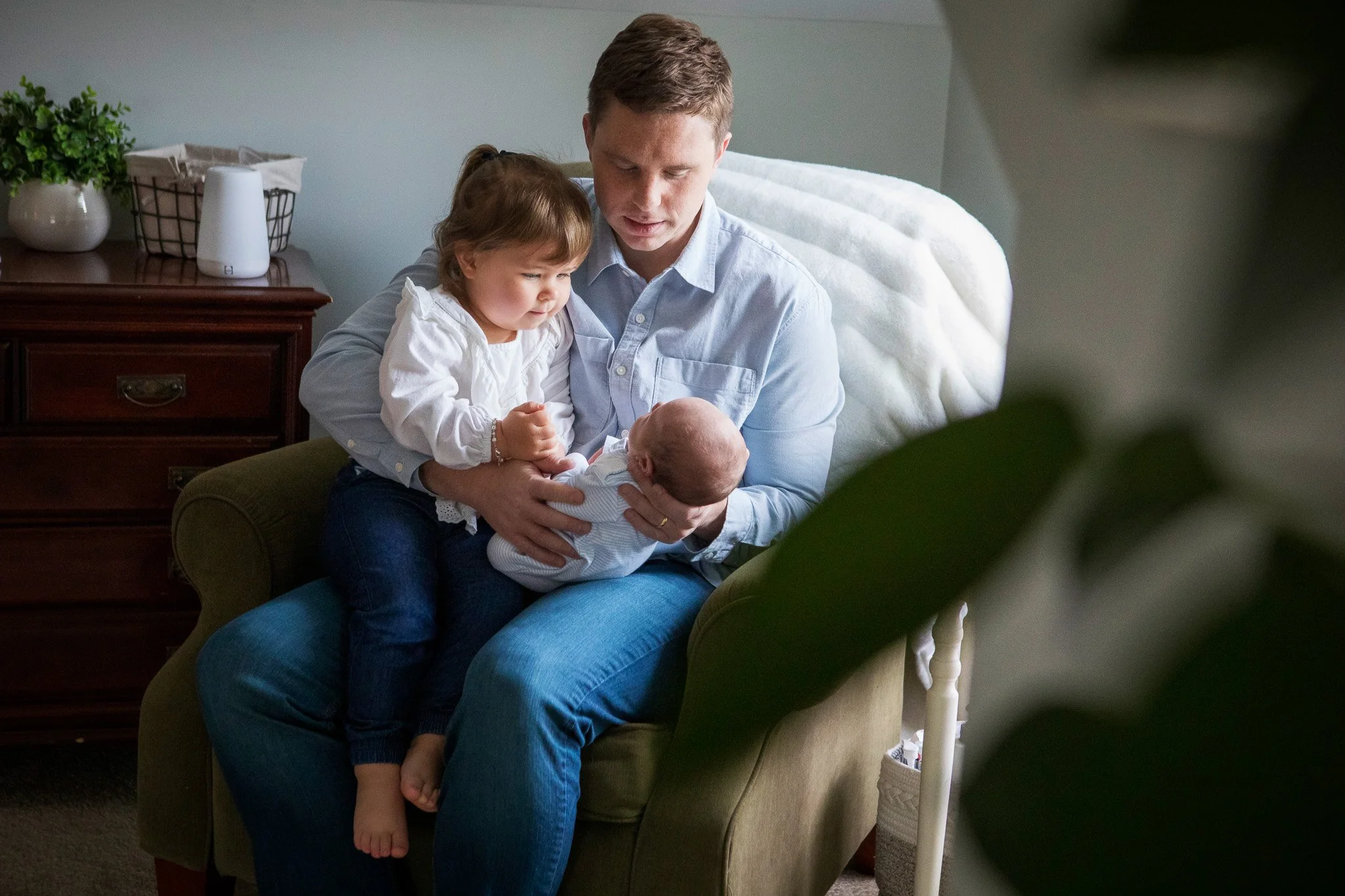 A family of three, with a young girl and a baby, sitting on an armchair in a cozy living room, sharing a tender moment.