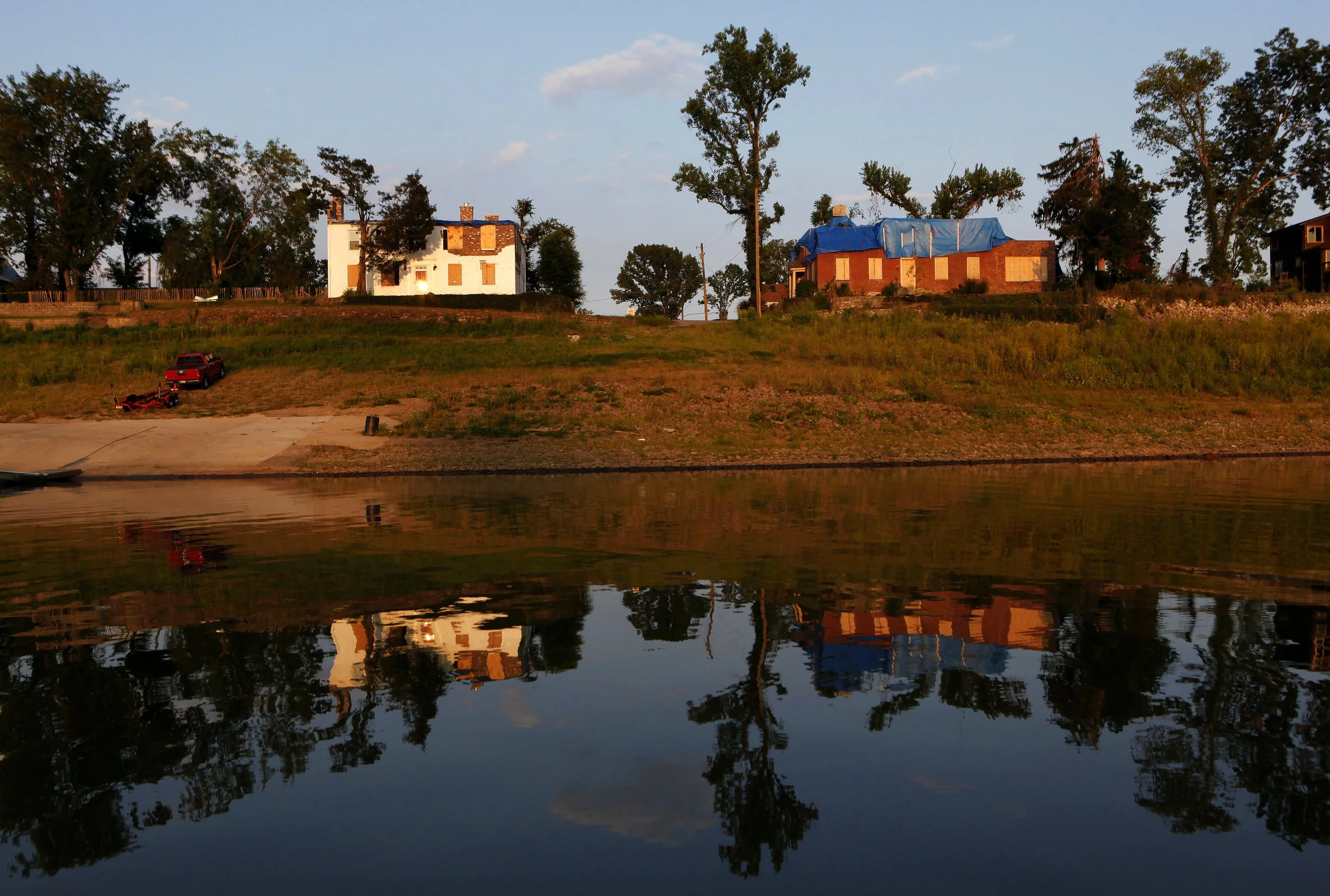 A lakeside view with two houses on a grassy hill, trees, a boat, and a red vehicle, all reflected in the water, during sunset.