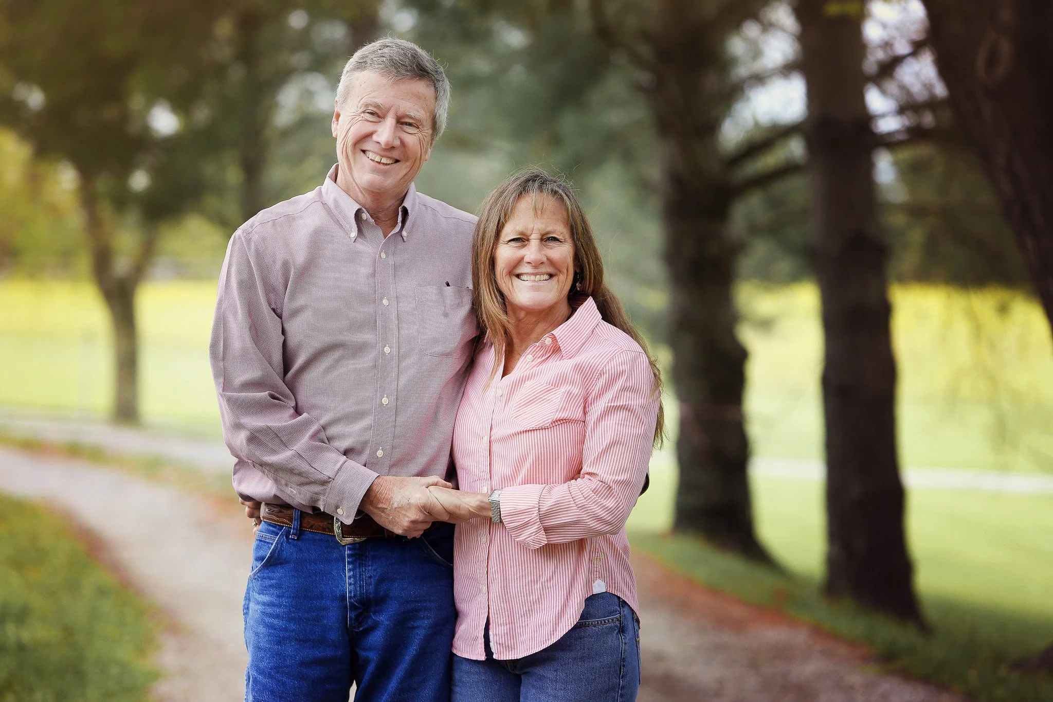 An elderly couple smiling and holding hands outdoors on a tree-lined path.