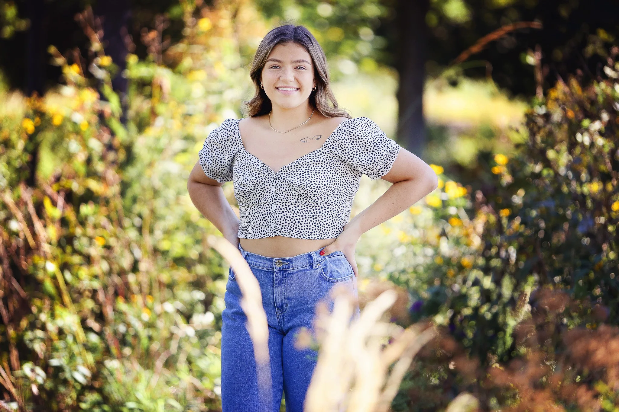 A young woman with brown hair smiling outdoors in a garden or park, wearing a white polka dot crop top and high-waisted blue jeans.
