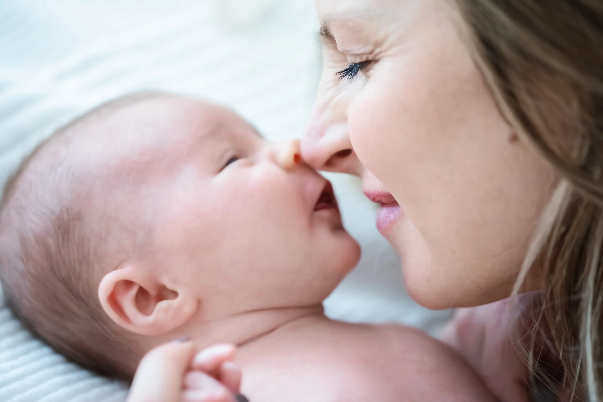 A mother and baby touching noses, close-up of faces, mom with closed eyes, baby smiling, both with light skin, lying on a soft white surface.