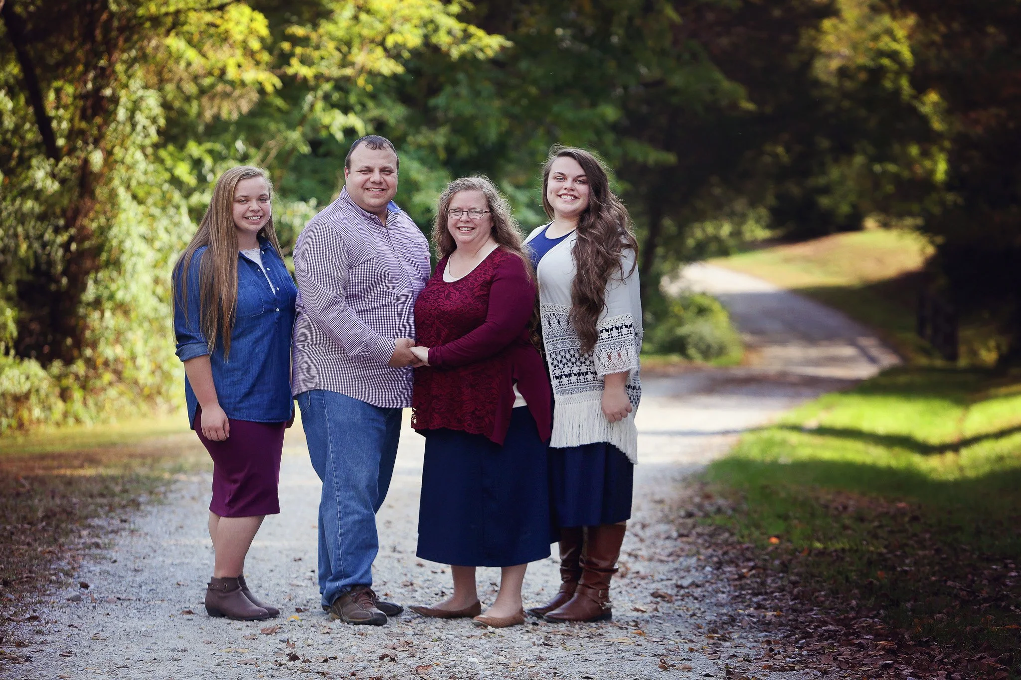 A family of four standing on a gravel path in a wooded park, holding hands and smiling at the camera.