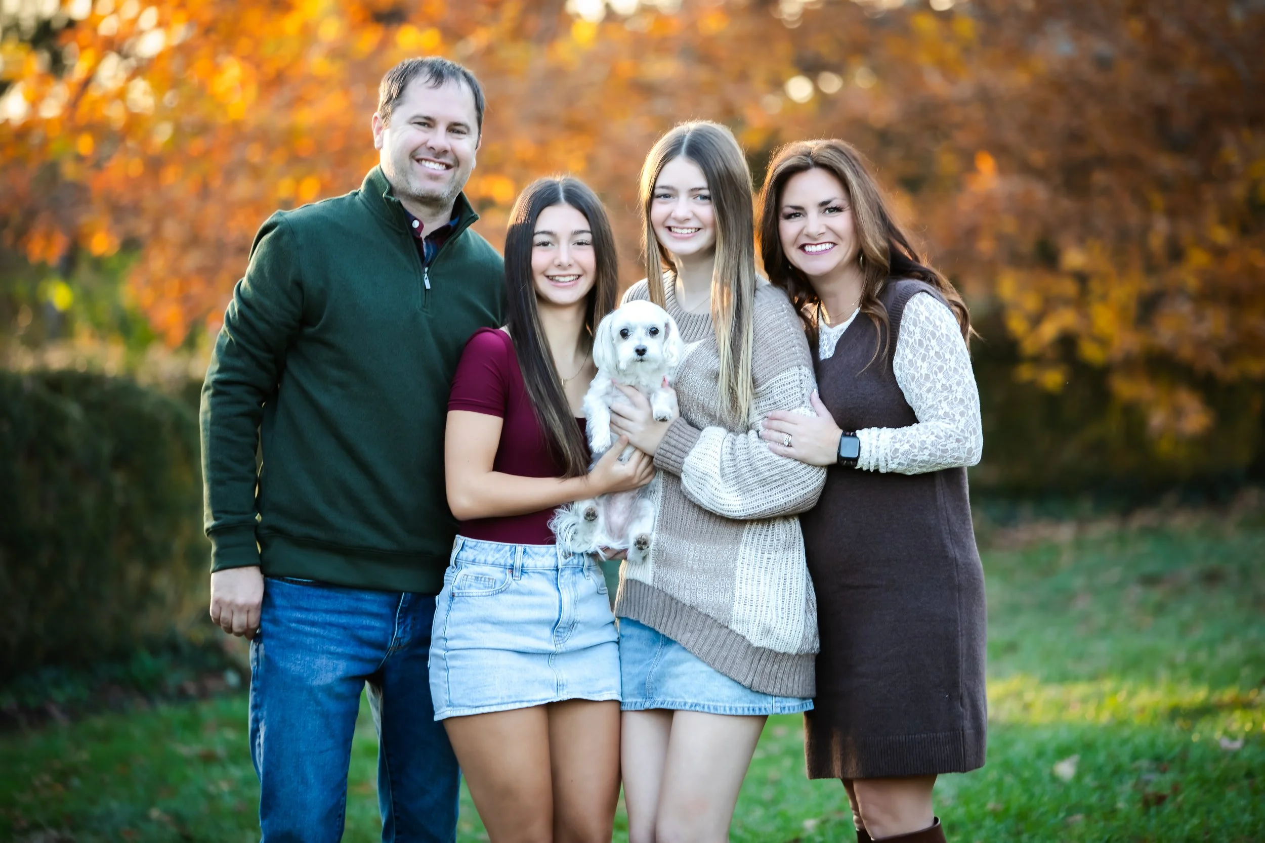A family of four with two women, a man, and a girl, holding a small white dog, standing outdoors during autumn with orange and yellow trees in the background.