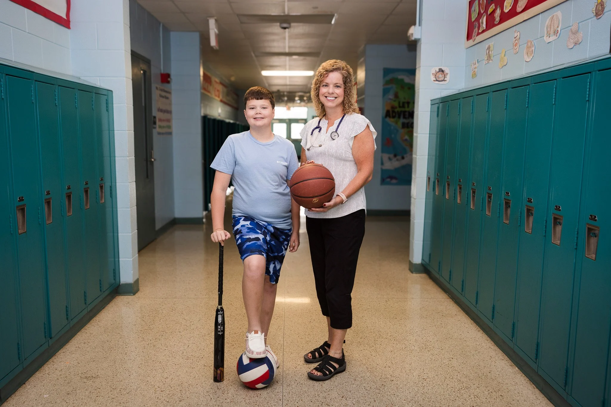 A boy with a cane and a volleyball standing next to a woman holding a basketball in a school hallway with lockers.