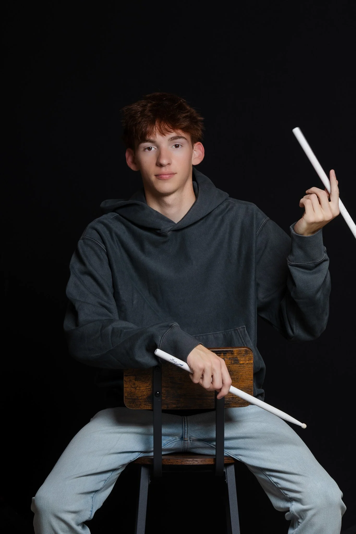 Teenage boy sitting on a wooden chair with black background, holding white sticks, wearing a dark gray hoodie and light gray jeans.