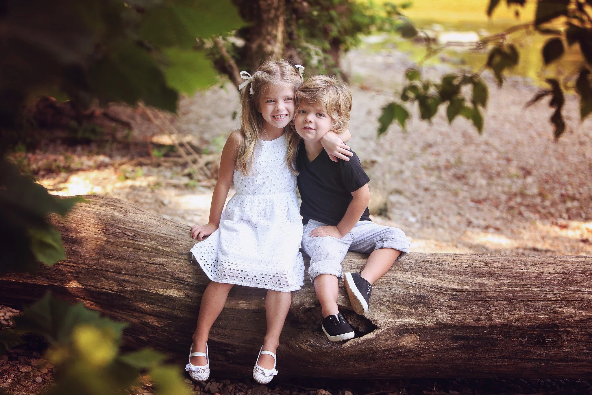 Two children, a girl in a white dress and a boy in a black T-shirt and rolled-up pants, sitting on a fallen tree trunk in a wooded area, embracing and smiling.