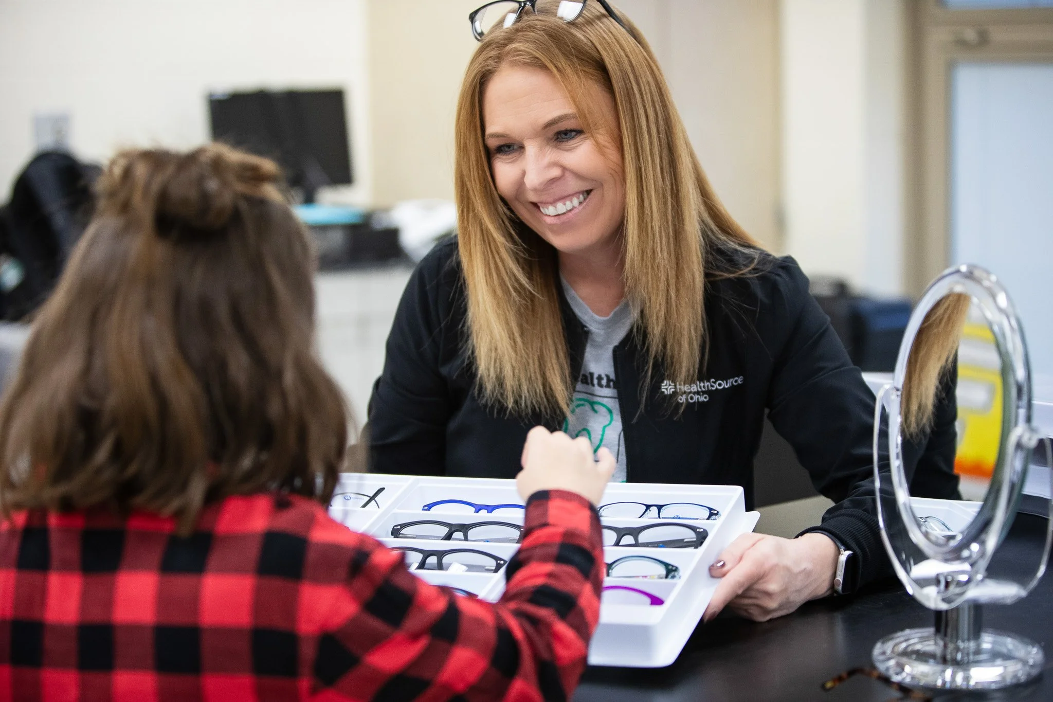 A woman with long red hair and glasses on her head is assisting a young girl in choosing glasses at an eye care center. They are sitting at a black table with a tray of various eyeglasses.