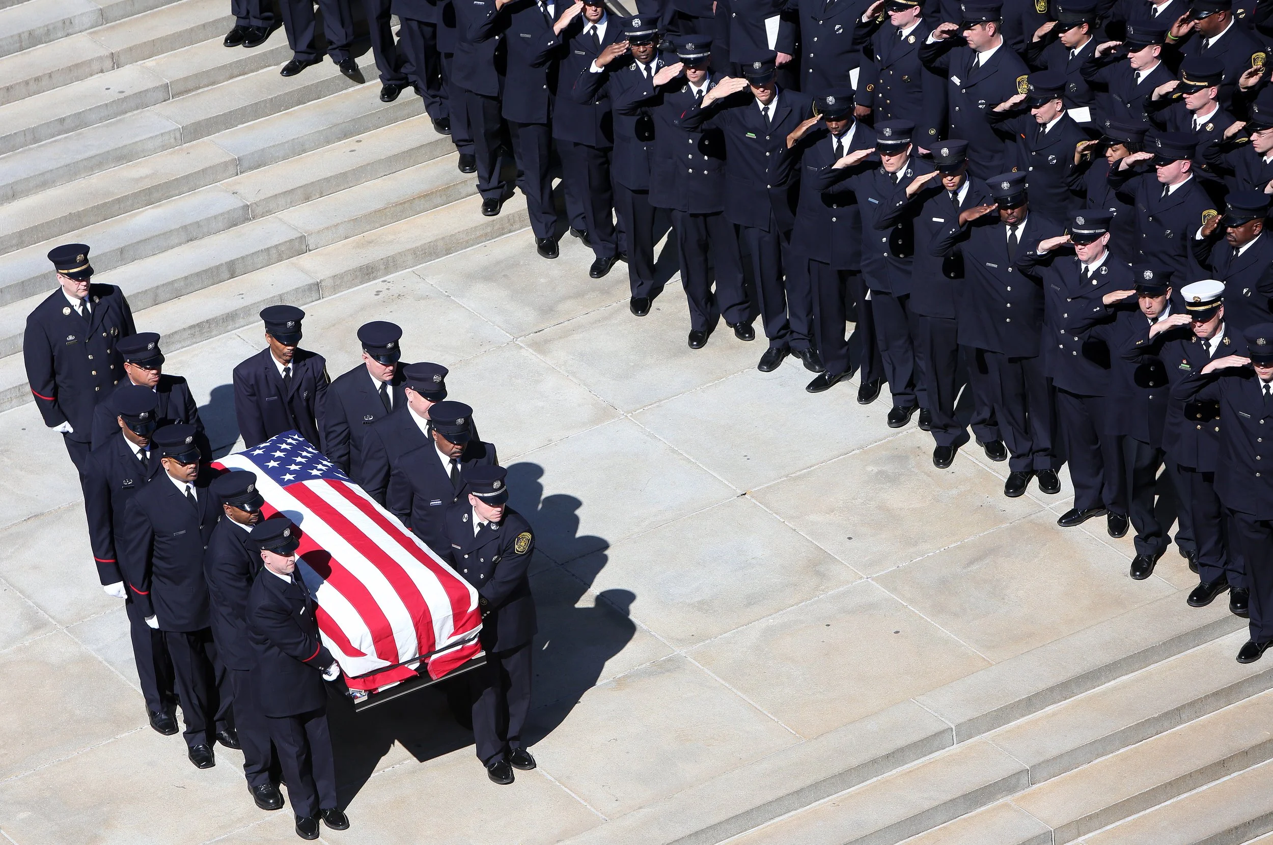 A military funeral procession with uniformed police officers carrying a casket draped with an American flag, saluted by a formation of officers on steps.