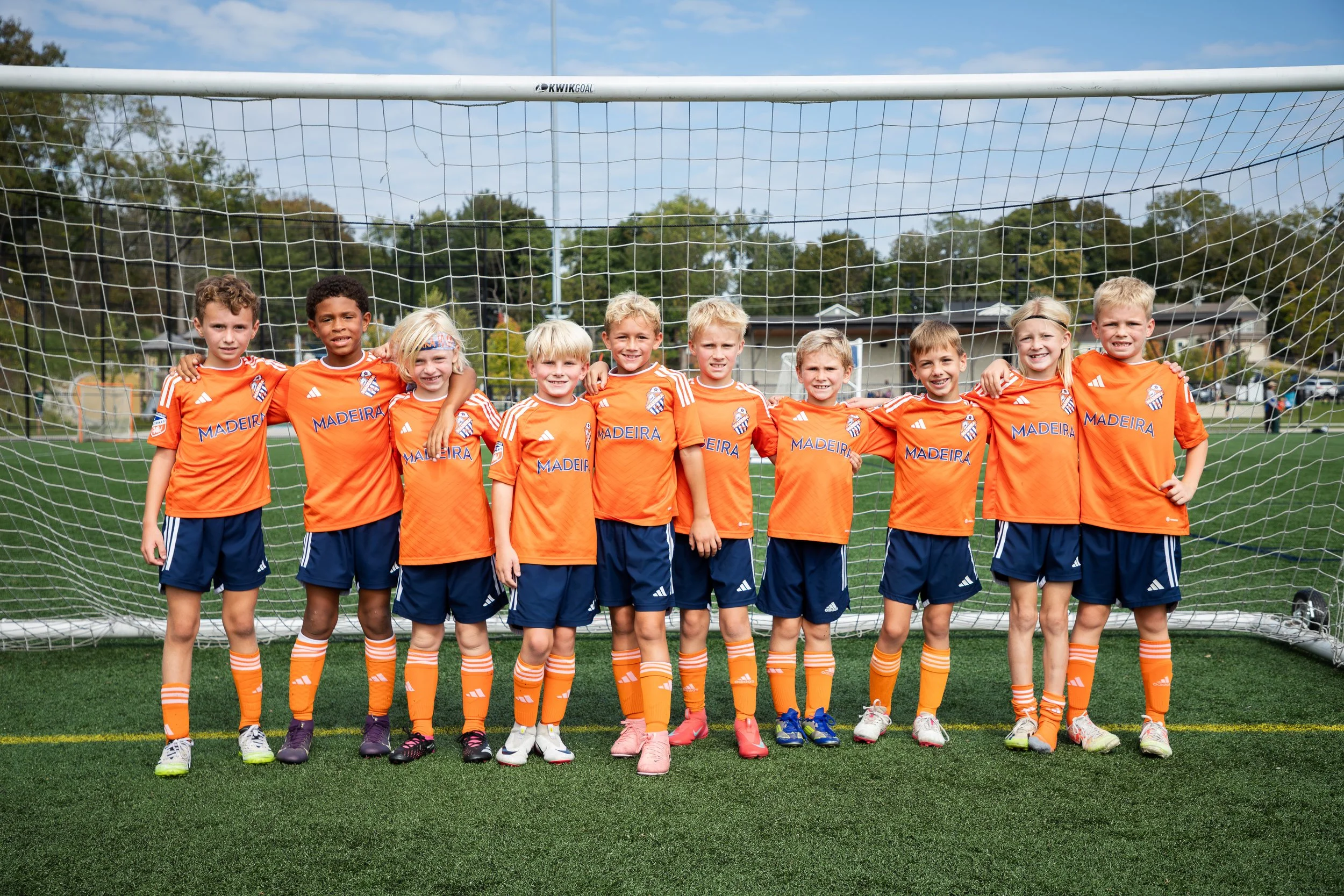 A youth soccer team of eleven kids dressed in orange jerseys and blue shorts, standing in front of a soccer goal on a field, smiling.
